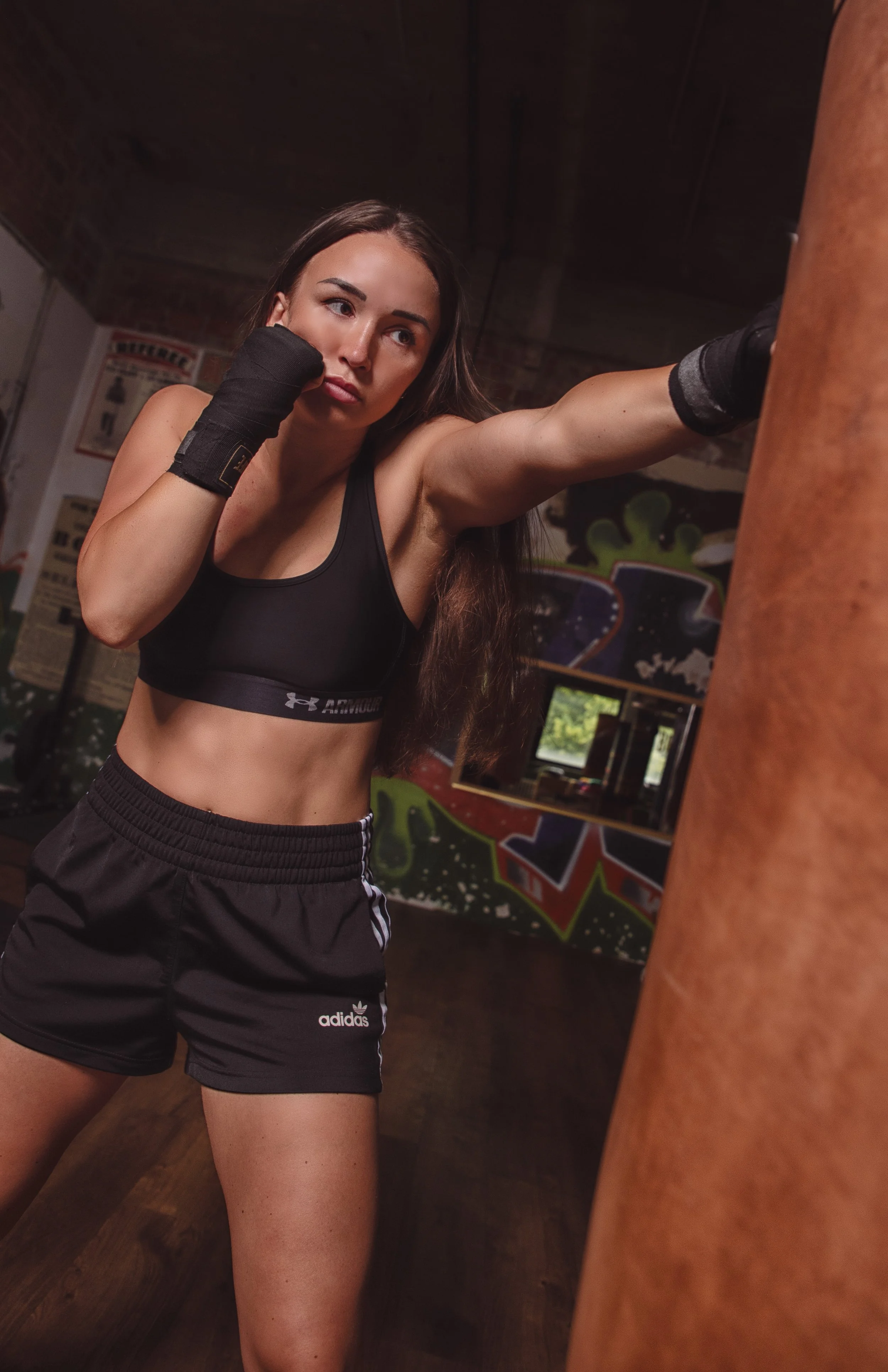 Frau beim Boxtraining in einem Fitnessstudio, trägt Sport-Bras und Shorts, steht vor einem Boxsack.