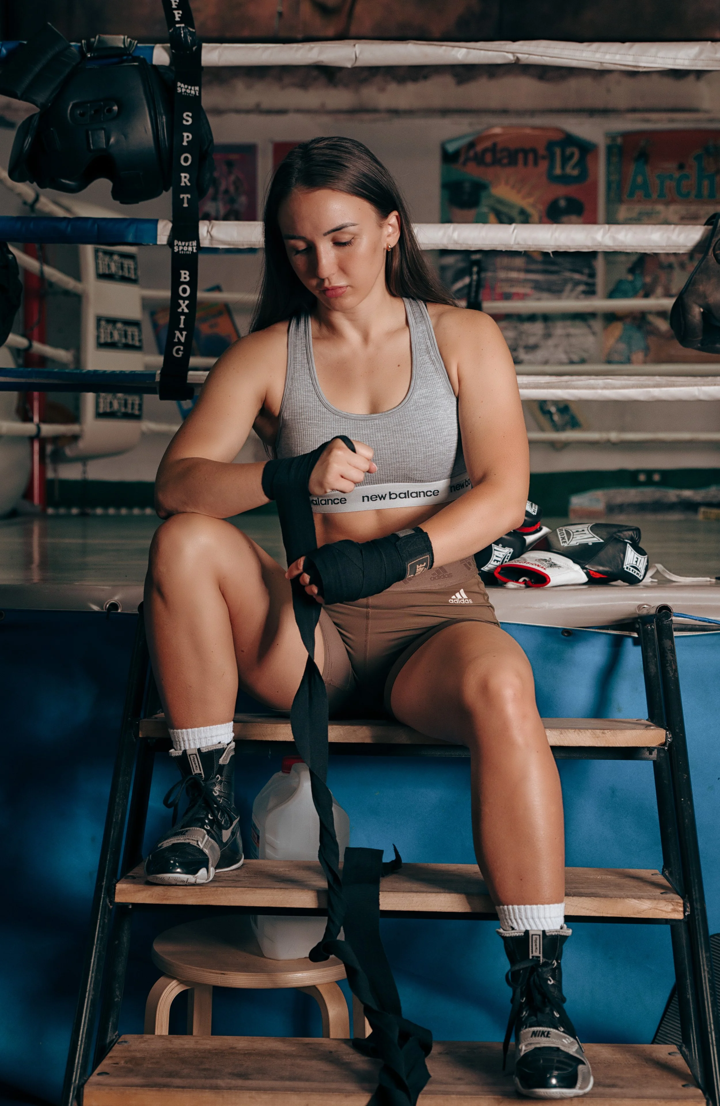 Eine junge Frau im Box-Trainingsraum sitzt auf einer Leiter und bindet ihre Boxhandschuhe. Sie trägt ein graues Sport-Bustier und braune Shorts. Im Hintergrund sieht man einen Boxring, Boxhandschuhe und Poster an der Wand.