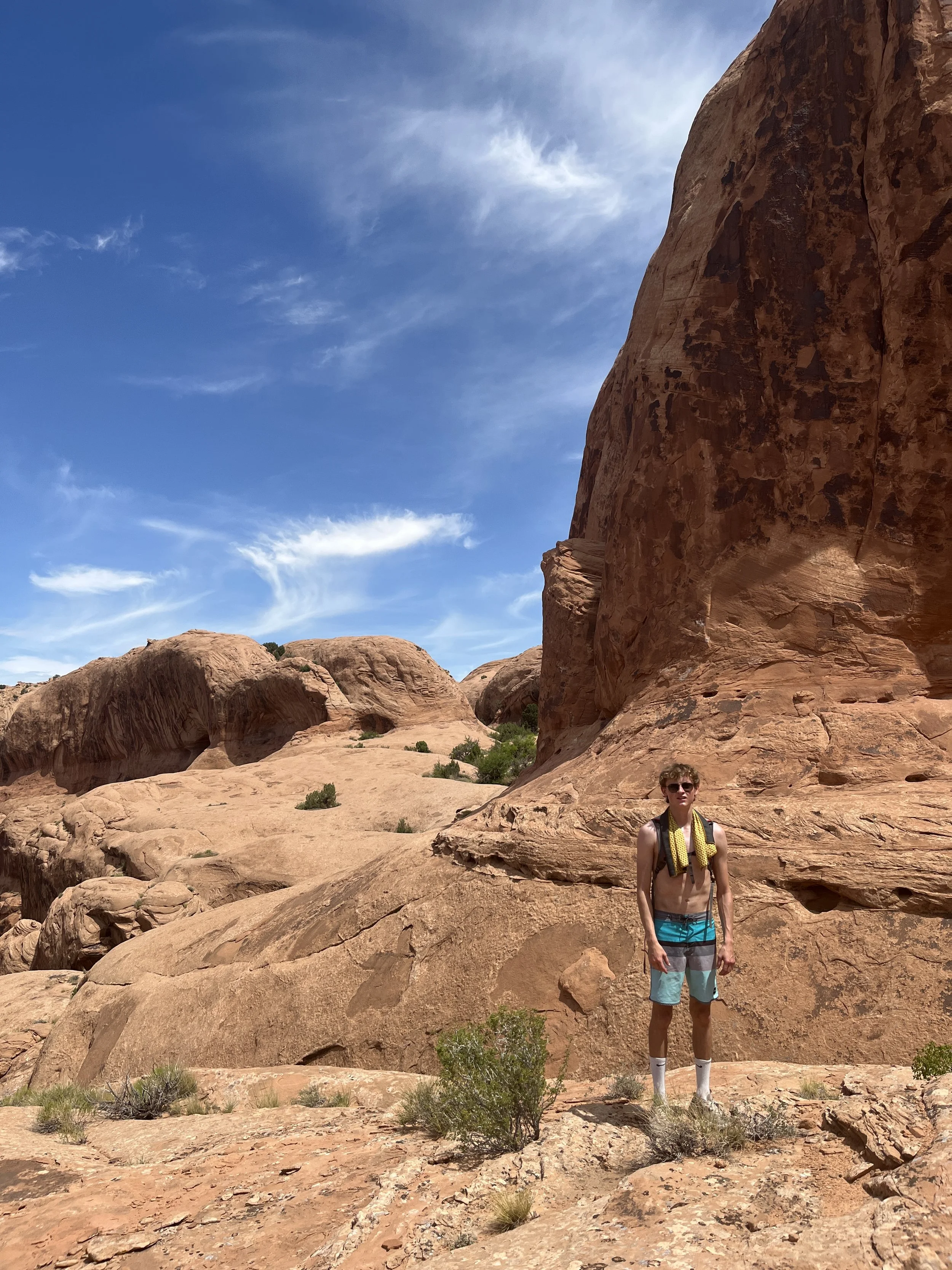A shirtless young man in blue and gray shorts with a yellow towel over his shoulders standing on red desert rocks under a partly cloudy blue sky.