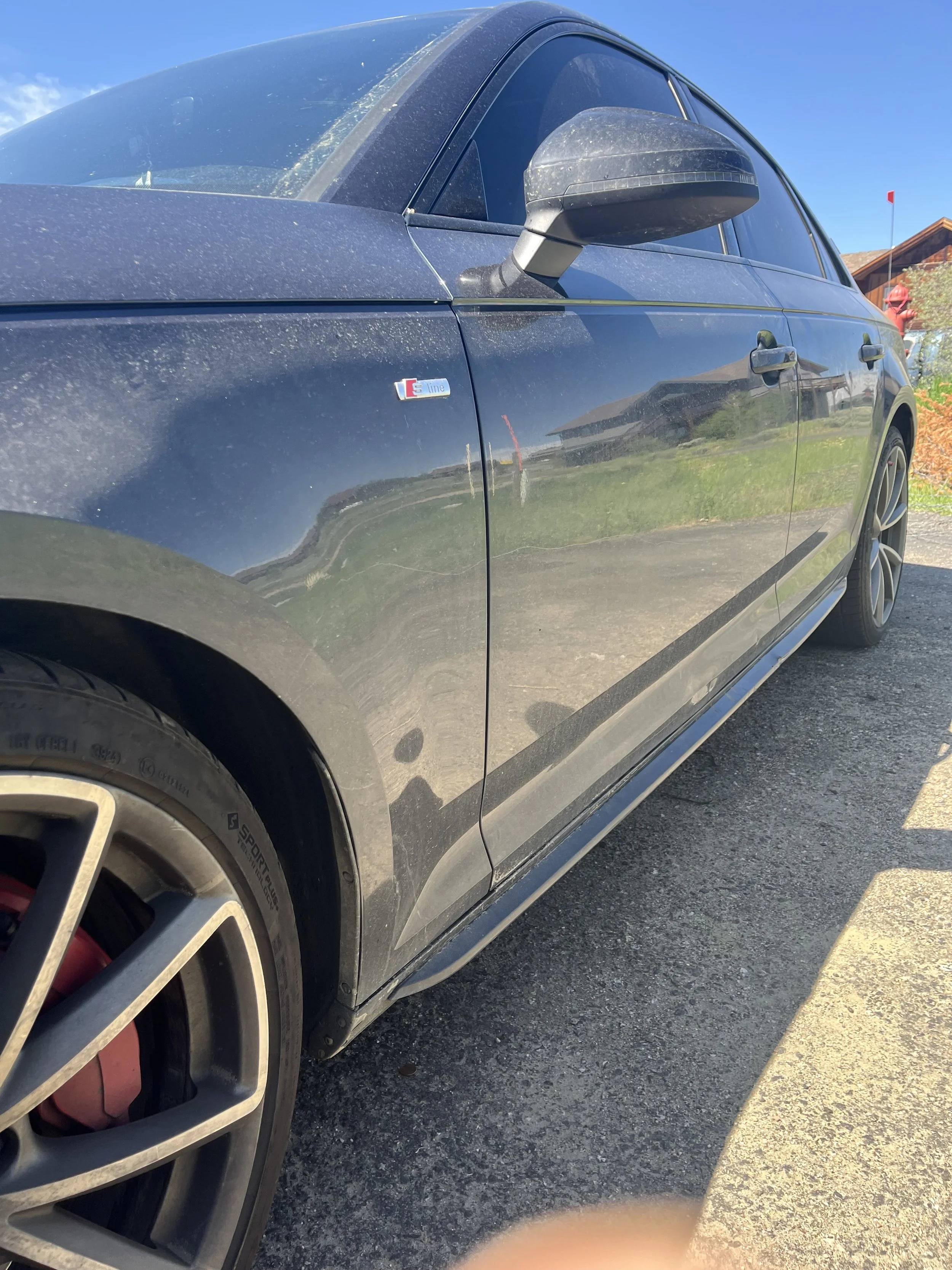 Black car with an 'S line' badge on the front fender, parked outdoors on a sunny day, with visible reflection on the side.