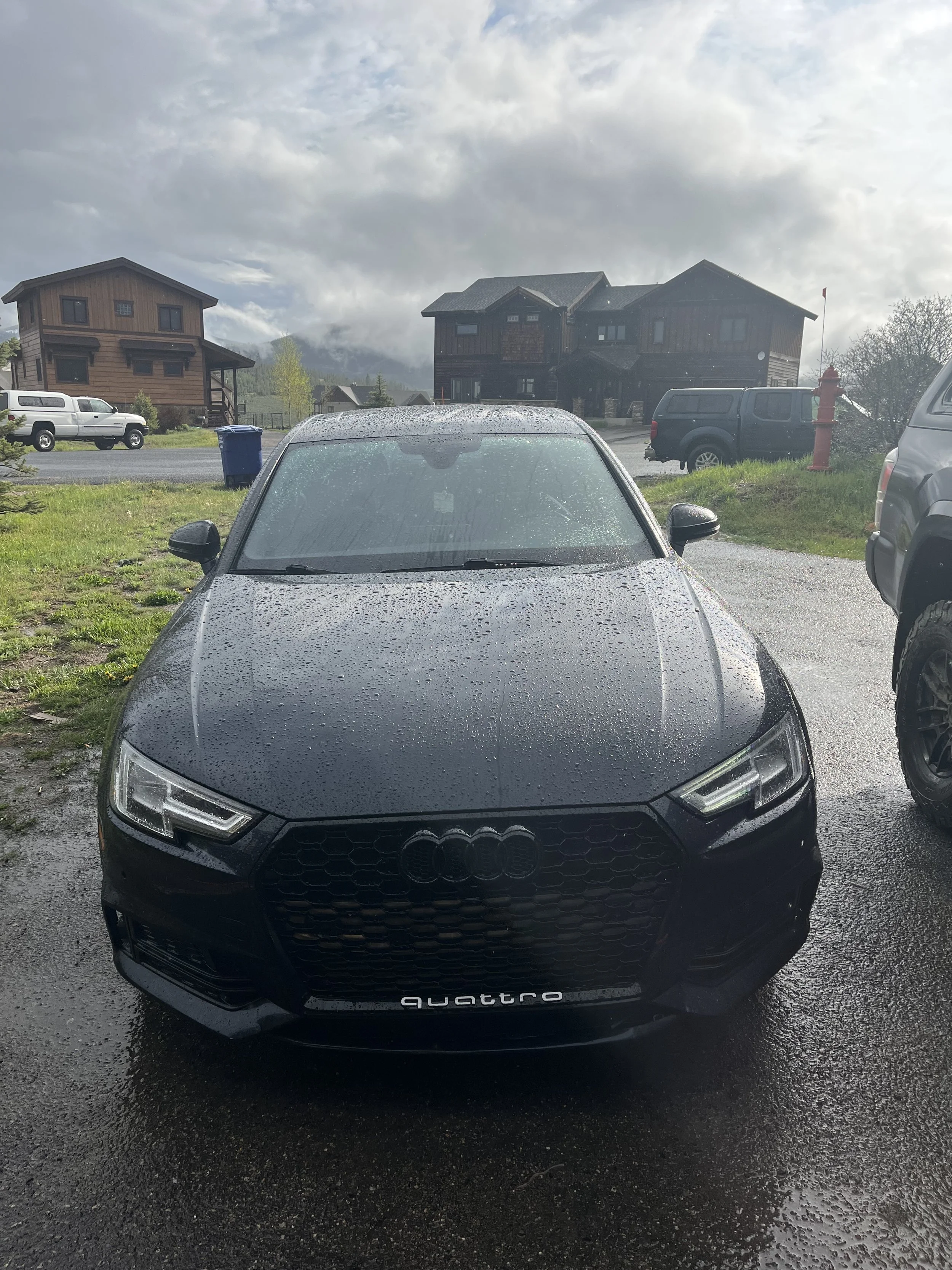 Black Audi Quattro parked outdoors on a rainy day, with water droplets on the windshield and hood, and houses and mountains in the background.