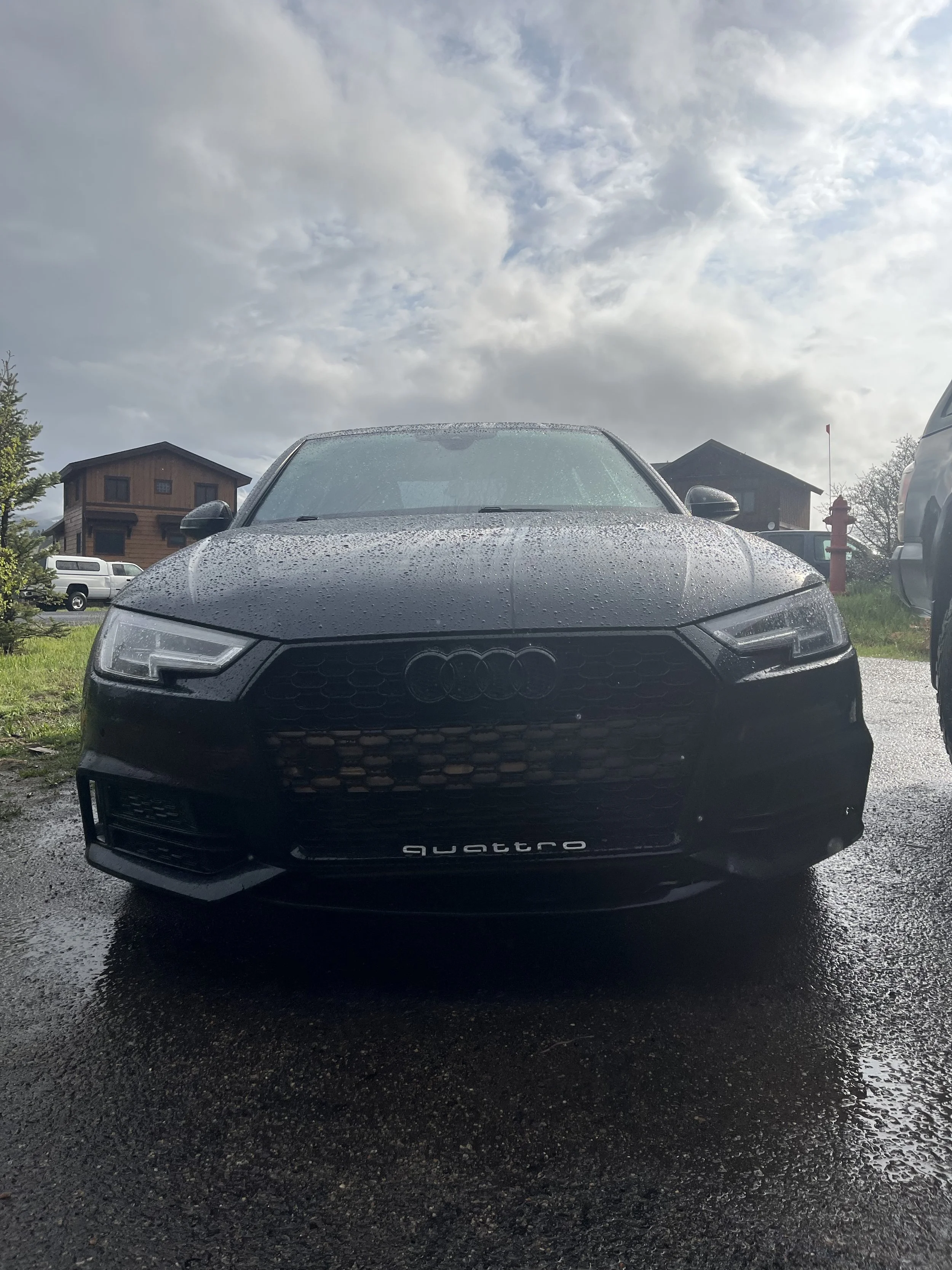 Front view of a black Audi Quattro car parked on a wet surface, with raindrops on the hood and windshield, under a cloudy sky.