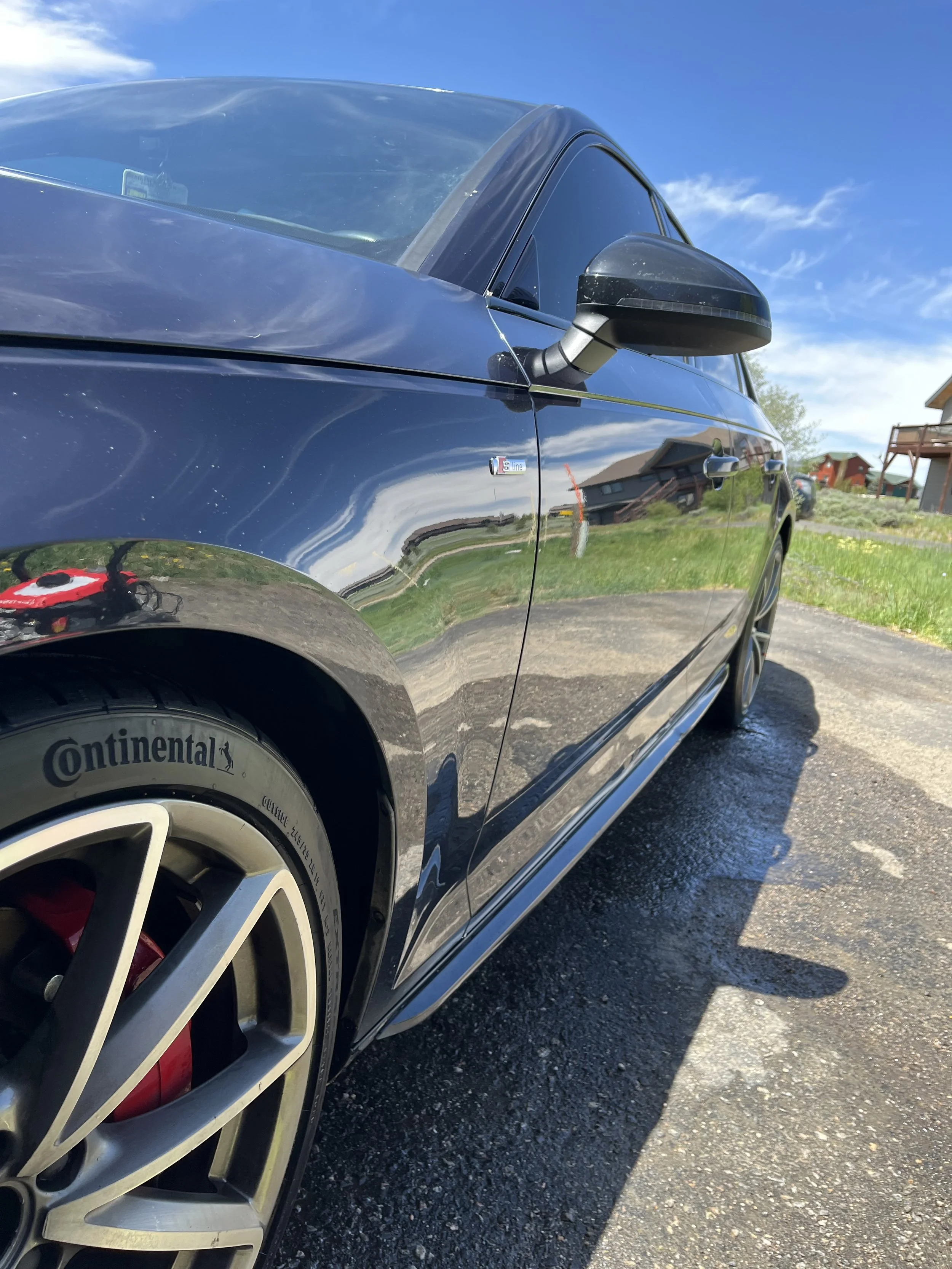 Close-up of a black sports car, showing the front left side, tire with Continental branding, and side mirror, parked outdoors on a sunny day with blue sky and houses in the background.