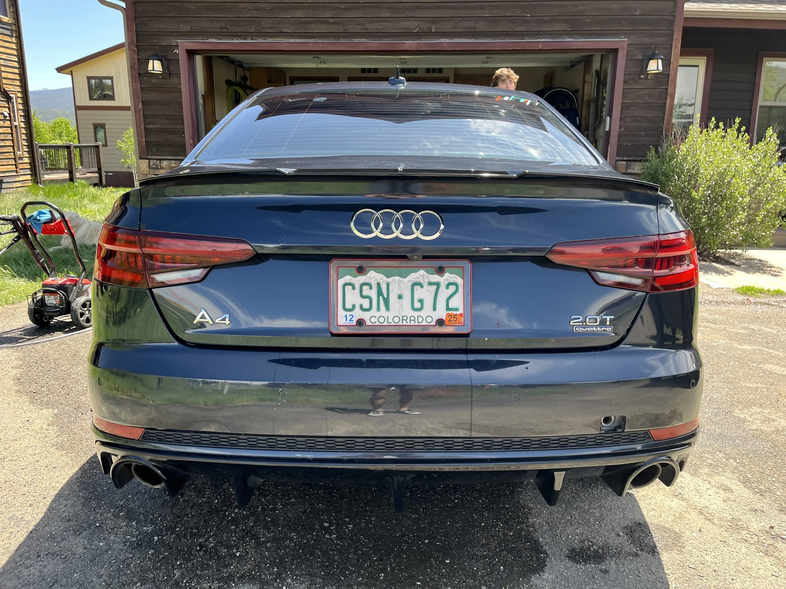 Rear view of a black Audi A4 with Colorado license plate CSN G72, parked in a driveway in front of a house, with a person partially visible inside the garage.