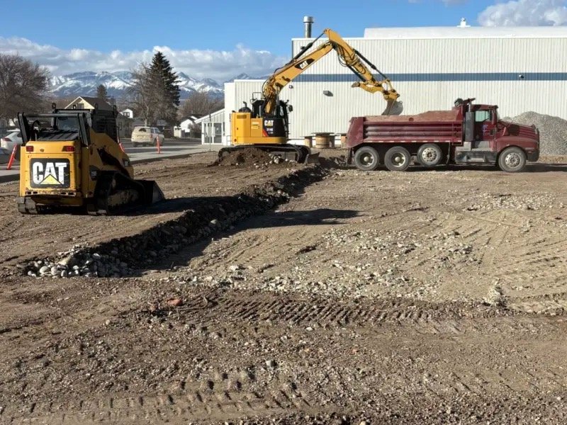 Construction site with a bulldozer, an excavator loading gravel into a red dump truck, and ground excavation work underway.