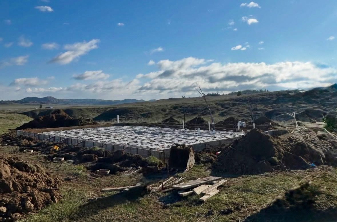 Construction site with concrete foundation and rebar, surrounded by dirt and construction materials, in a rural landscape with hills and a blue sky with clouds.