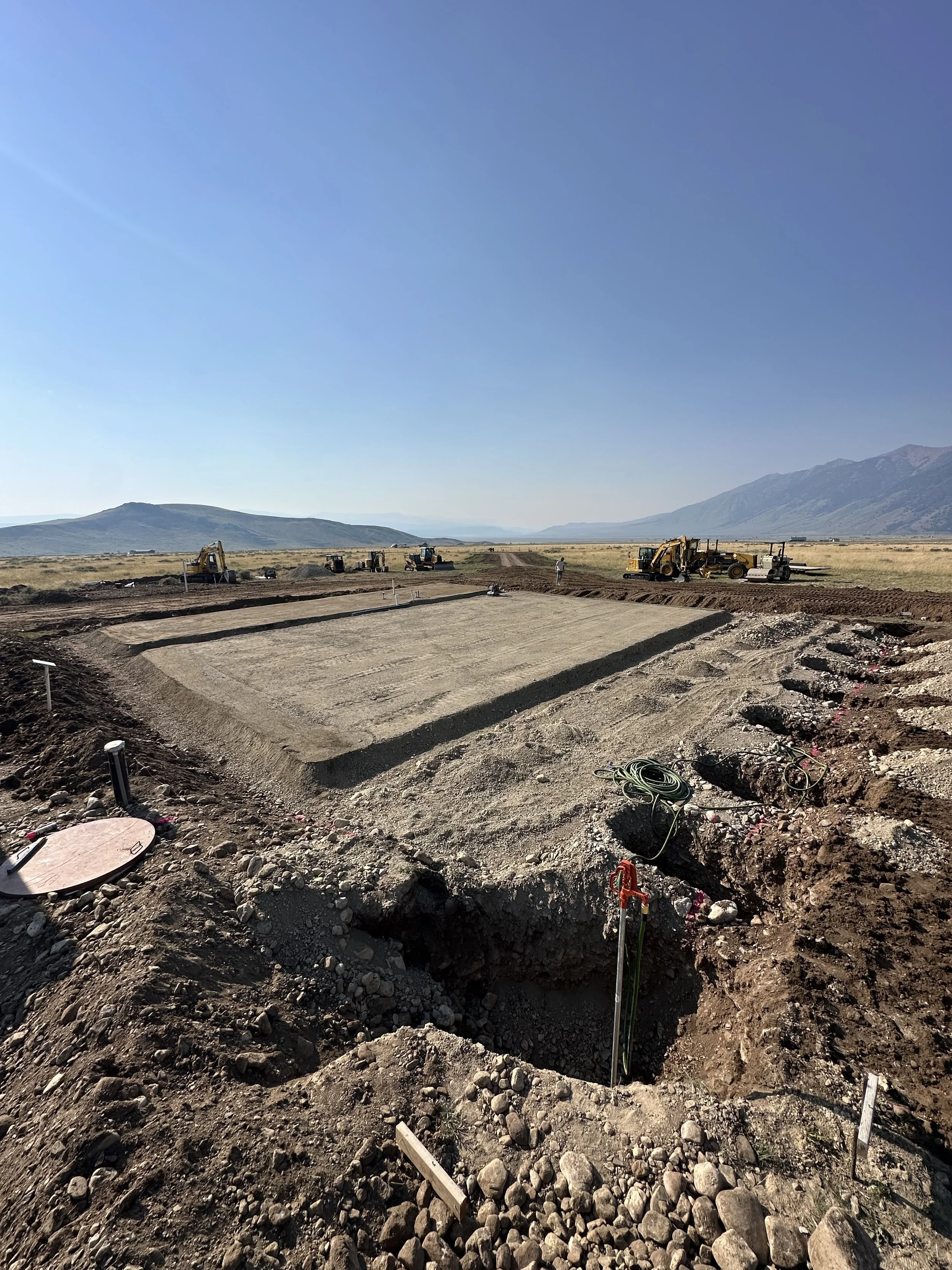 Construction site with excavation, concrete work, and machinery in a rural area with mountains in the background.