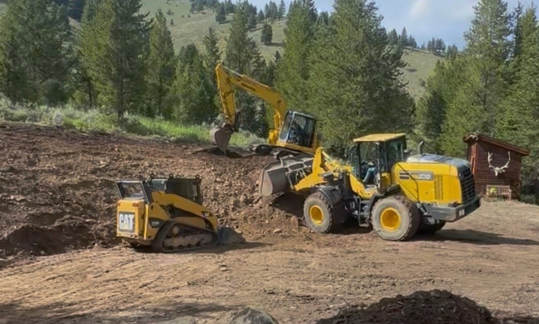 A construction site in a forested area with two yellow excavator machines working on the dirt slope.