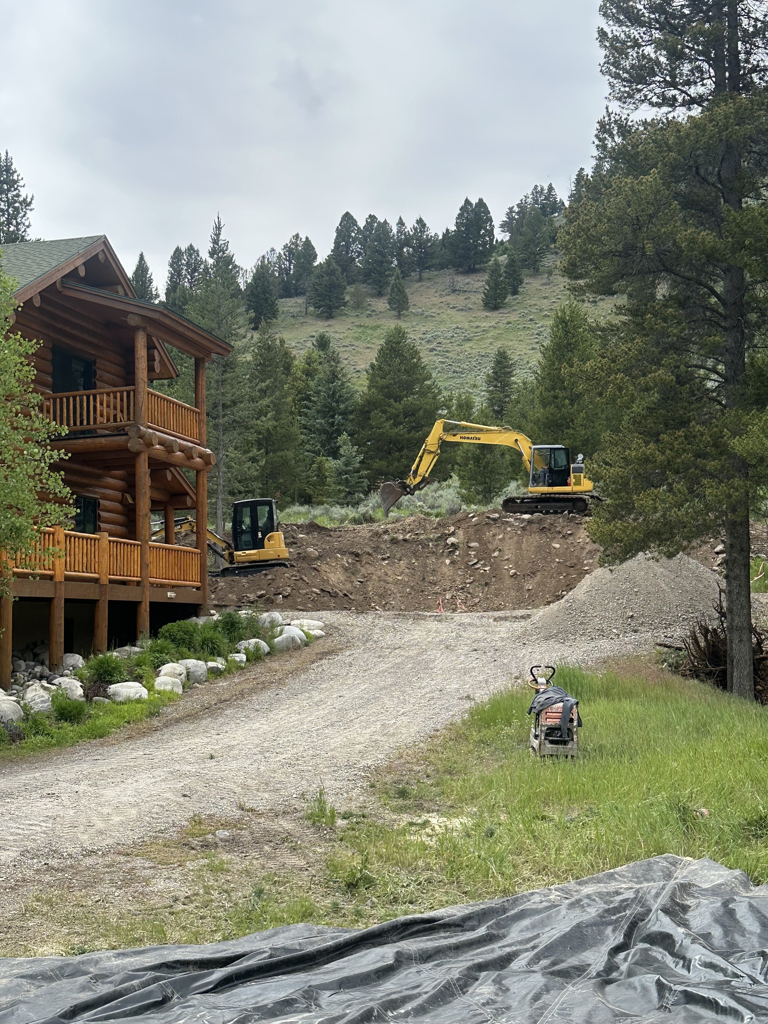 Construction site in a wooded area with trees and a mountain in the background. Two yellow excavators are working, one on the right side of the scene and the other partially visible on the left. To the left, a wooden log cabin with a balcony is visible. The ground is partly dug up, with a pile of gravel and a black tarp covering part of the foreground. A small lawn mower and some rocks are also visible.