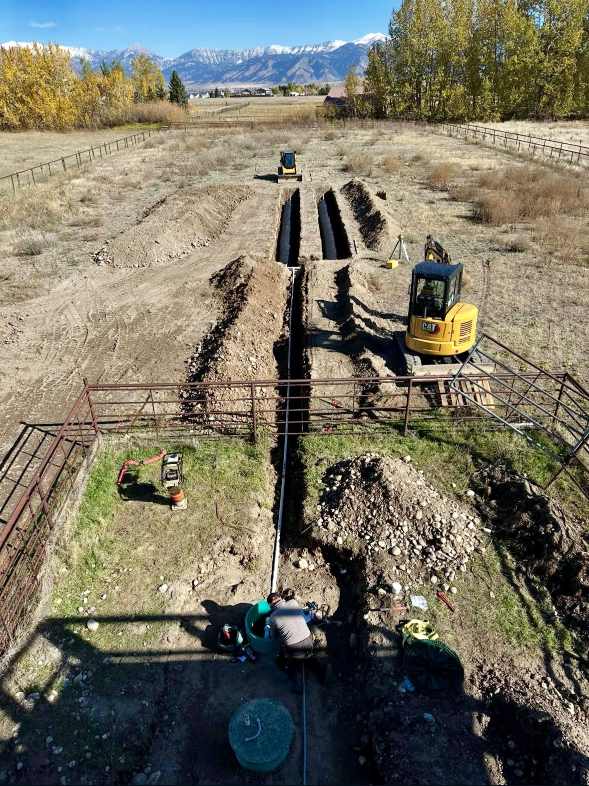 Construction workers installing a pipeline trench on a farm field with mountains in the background, using excavators, and various construction tools and materials.