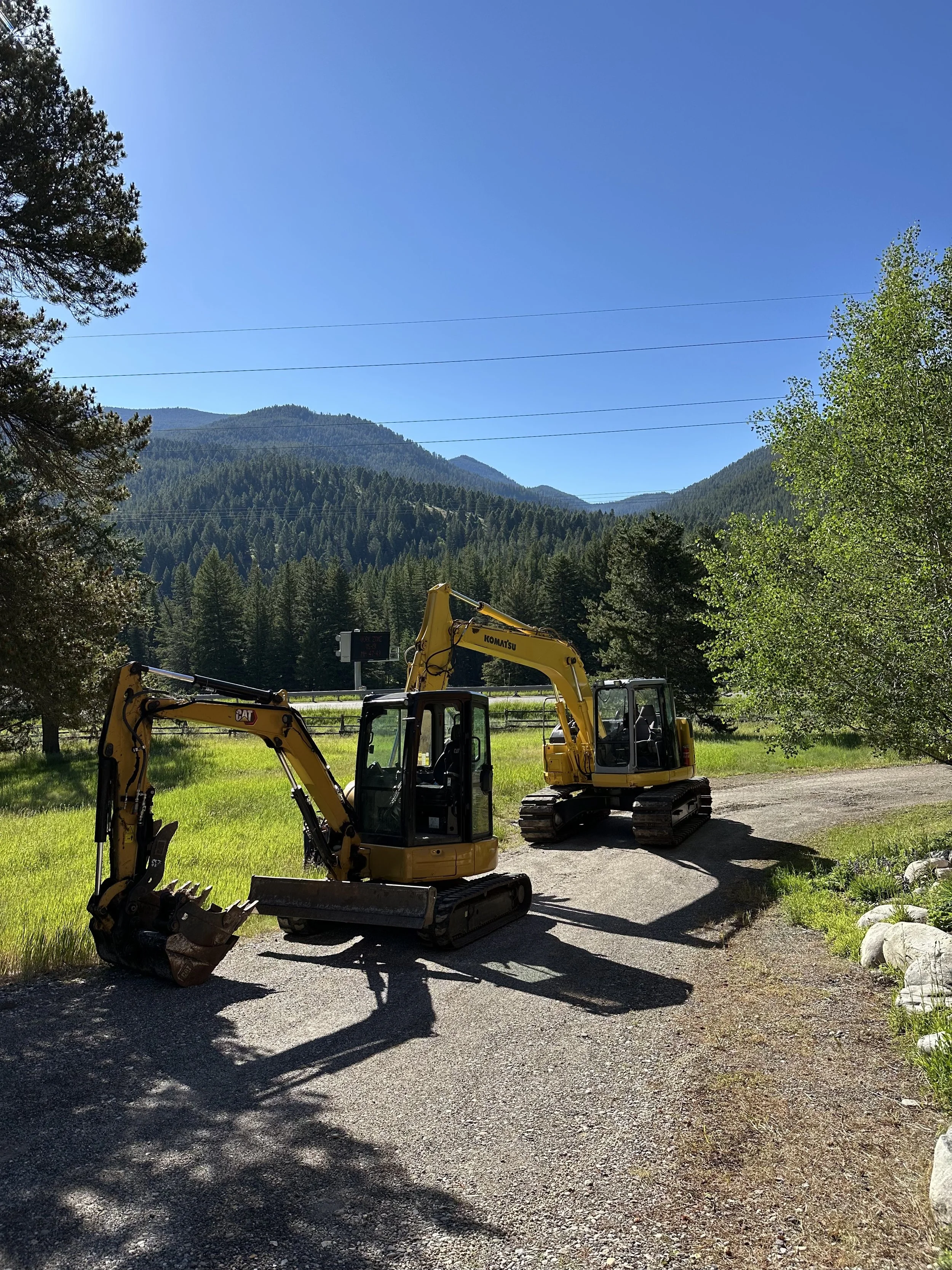 Two yellow excavators parked on a gravel path with lush green grass and trees, mountains in the background, and a clear blue sky overhead.