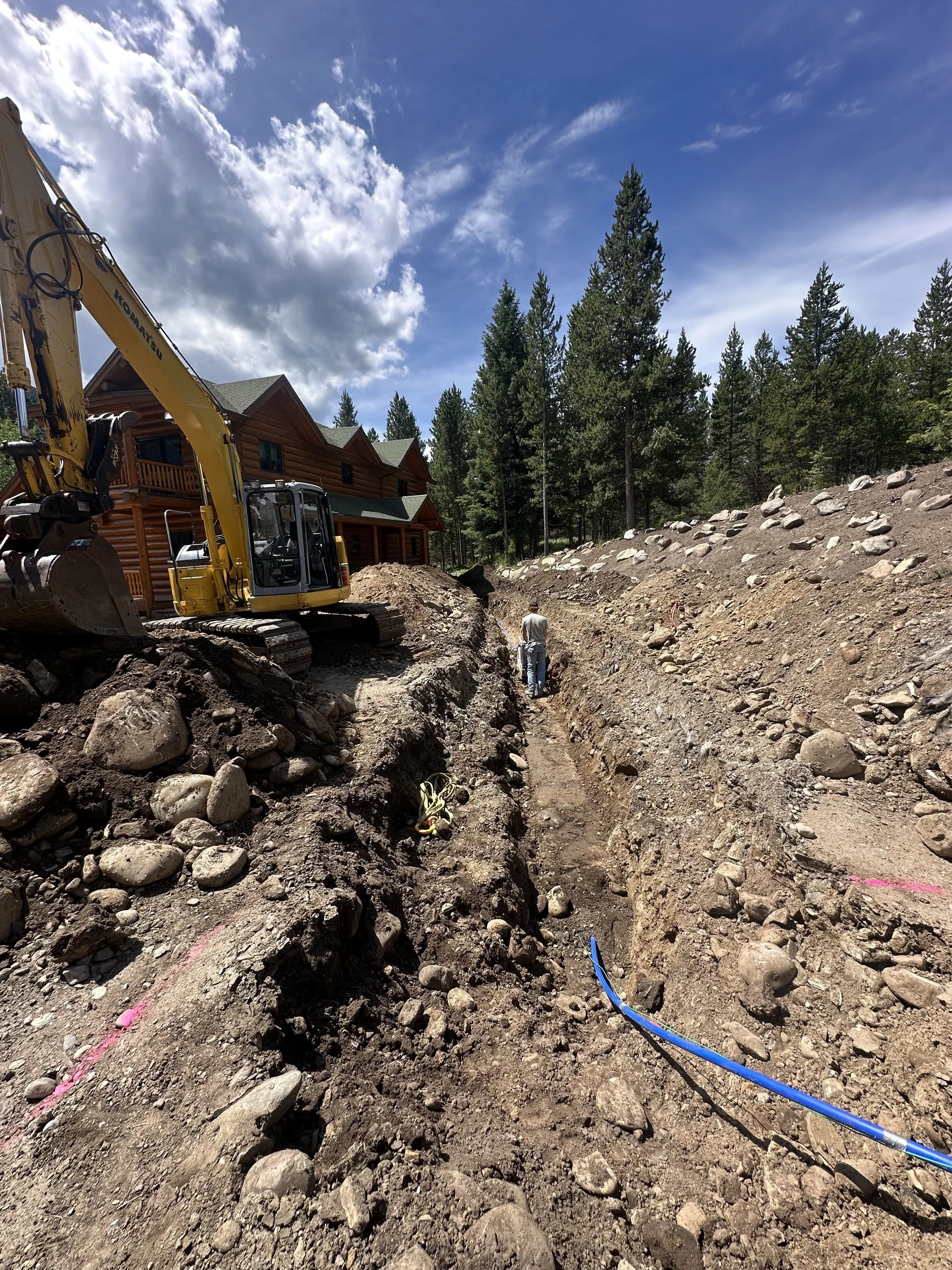 Construction site with a yellow excavator digging a trench, a man standing in the trench, a log cabin, and tall pine trees in the background under a partly cloudy sky.