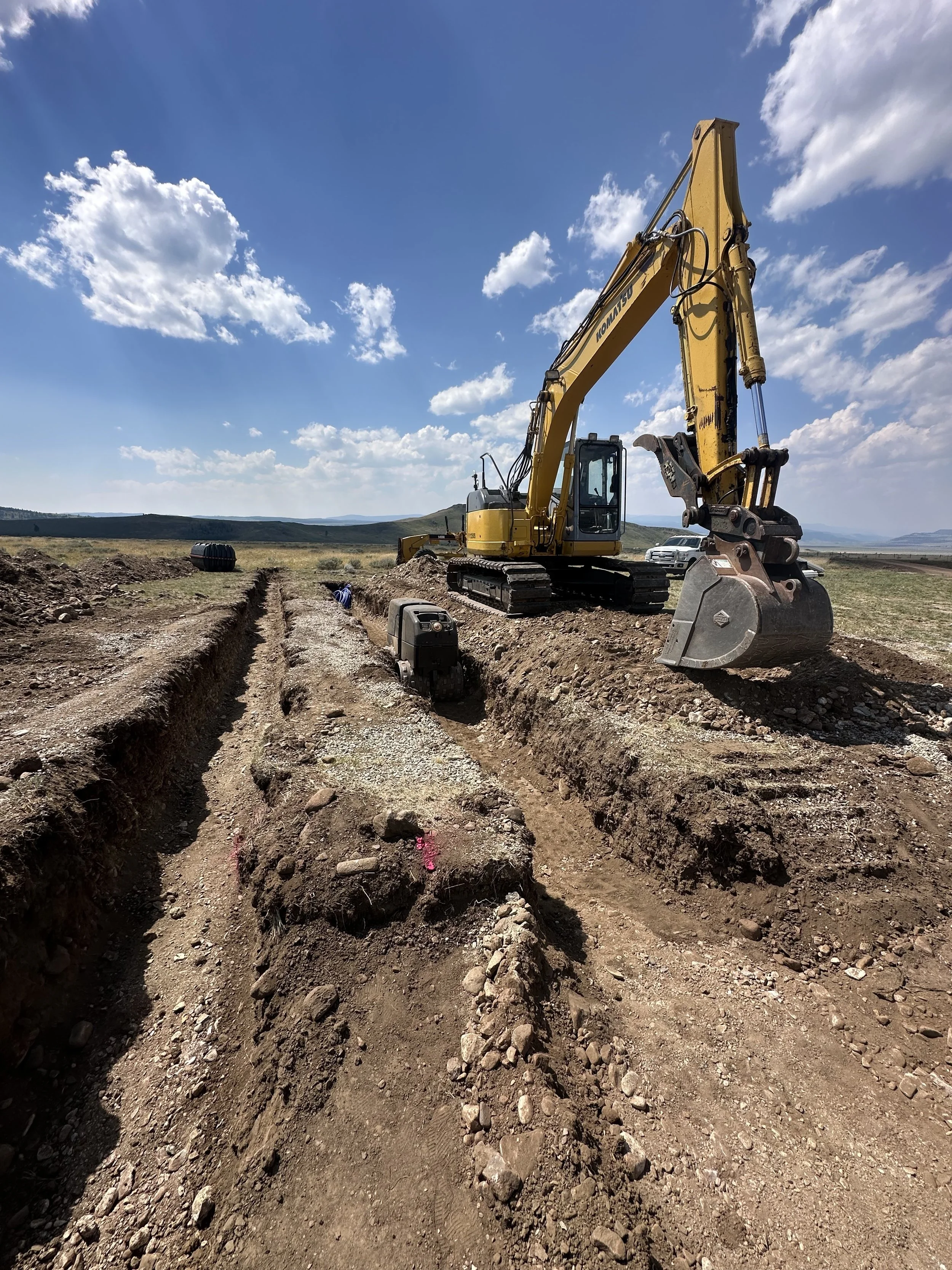 Construction site with an excavator digging a trench in a rural area under a partly cloudy blue sky.