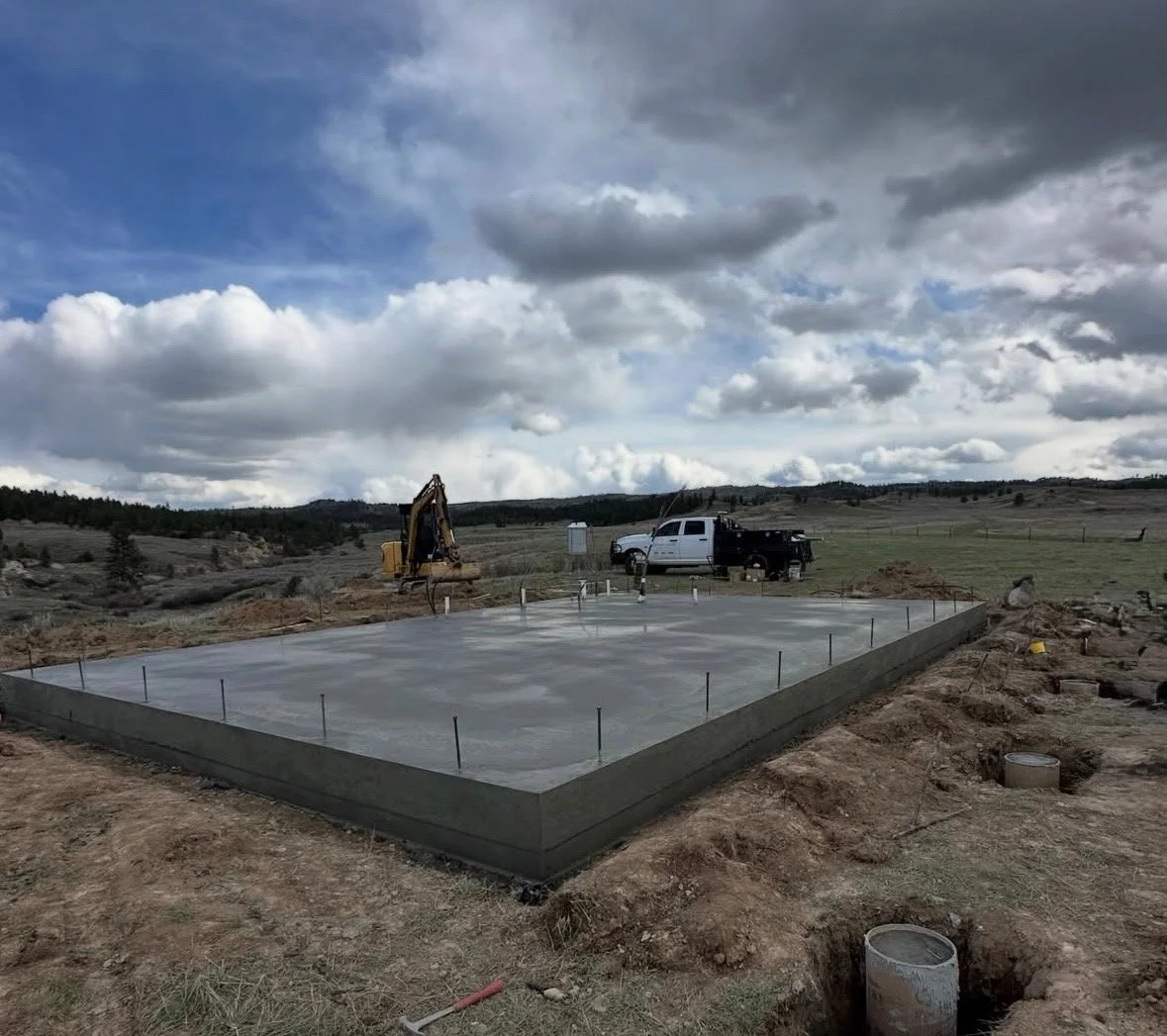 Concrete foundation being poured on a construction site with a pickup truck and construction equipment in an open landscape under a cloudy sky.