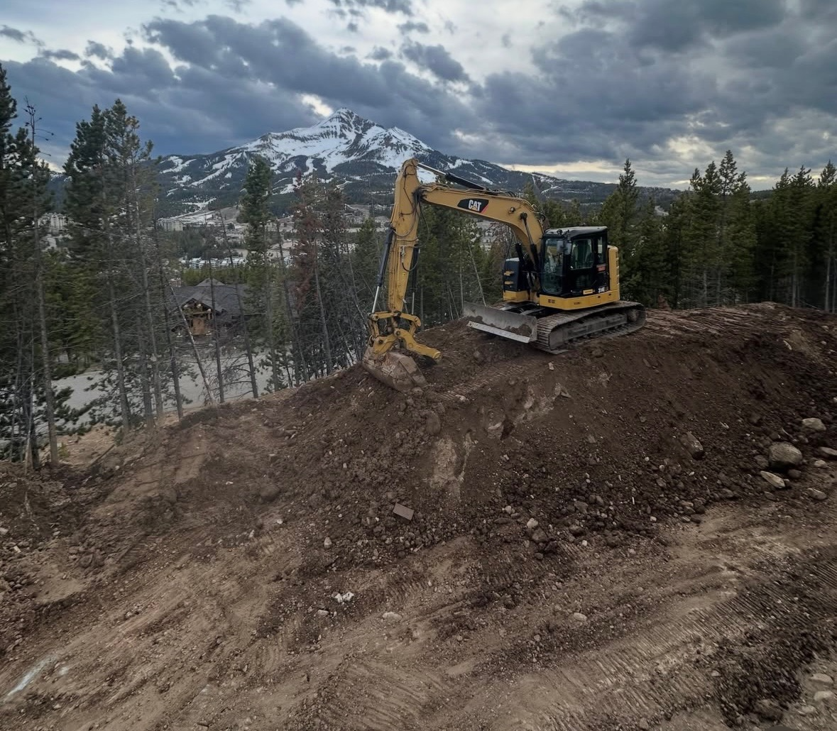 A yellow CAT excavator working on a dirt slope with a mountainous backdrop, trees, and a cloudy sky.