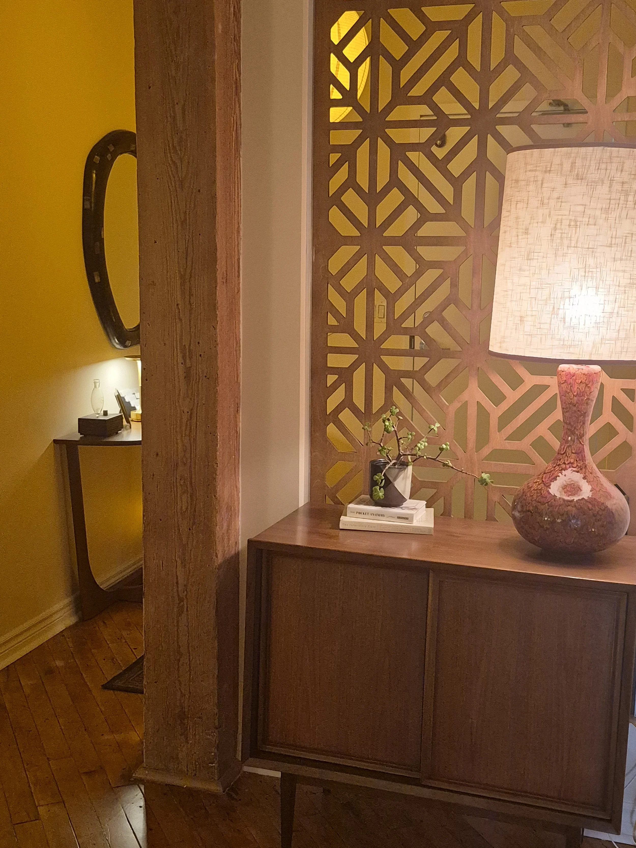 Interior of a room with a wooden sideboard, a decorative screen, a table lamp with a textured shade, a small potted plant, and books. There is a wooden floor and yellow wall.