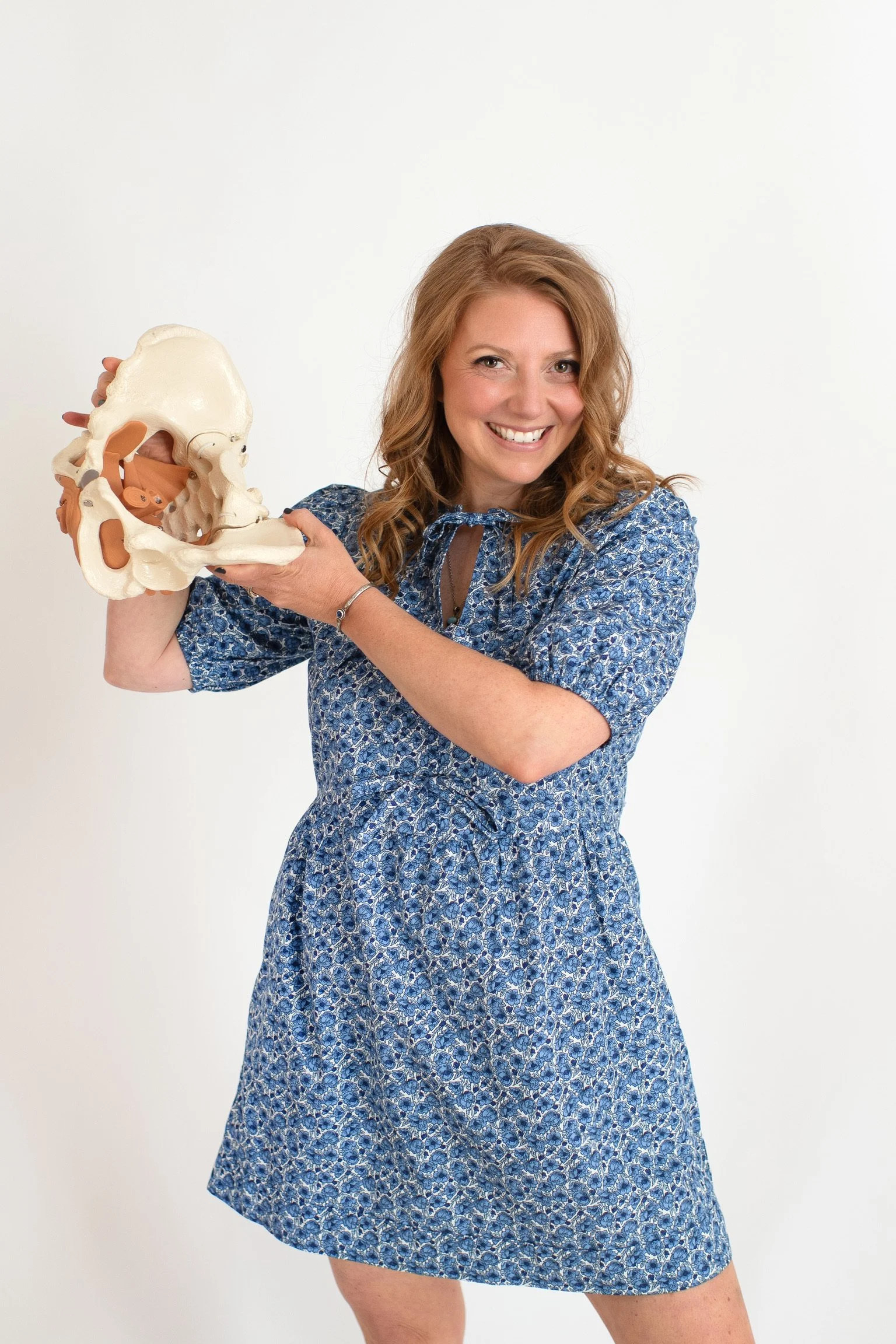 Dr. Krystal Couture of The Pelvic Acu, pelvic acupuncturist, with curly red hair smiling while holding a pelvic anatomical model against a plain white background.