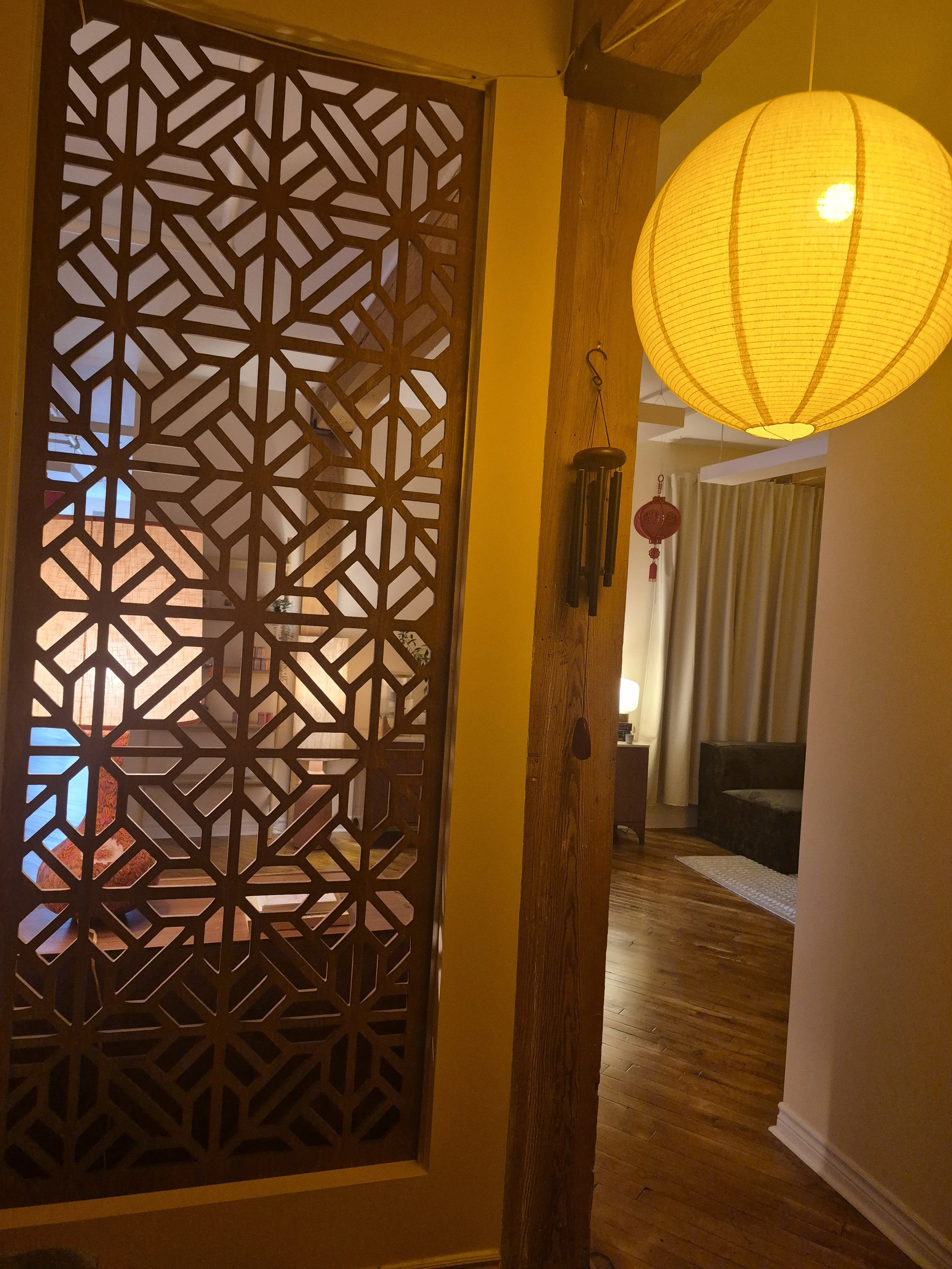 Interior view of a room with a decorative wooden partition, a yellow hanging lantern light, a wind chime, and hardwood floors.