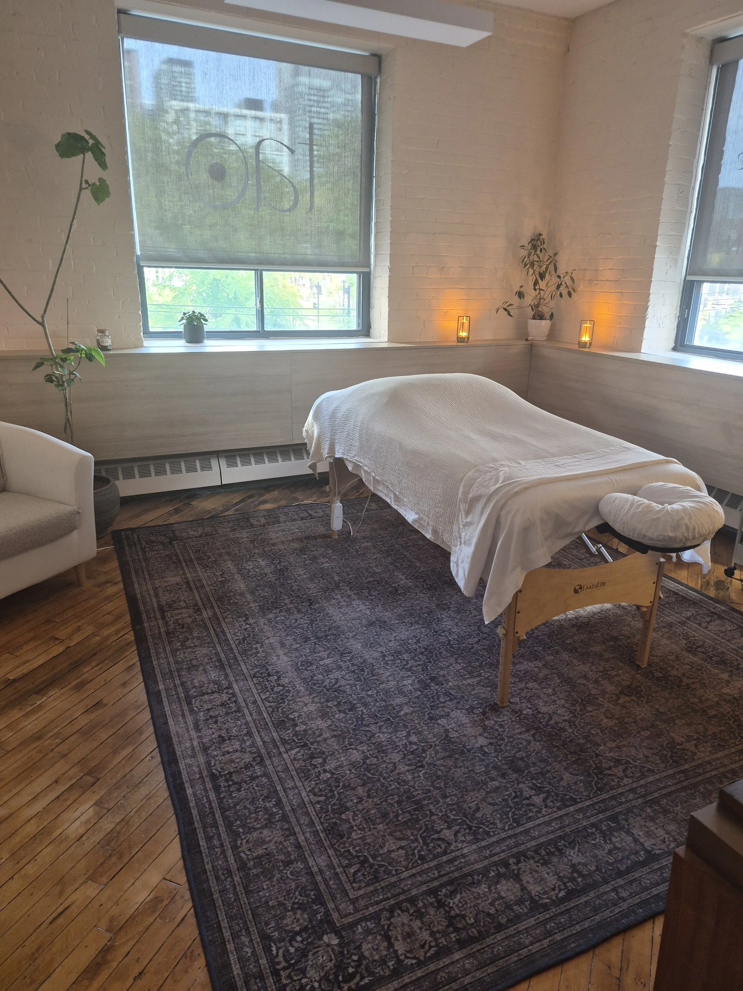 A Treatment table covered with a white sheet in a room with large windows, plants, candles for ambient lighting, a wooden floor, and a rug.