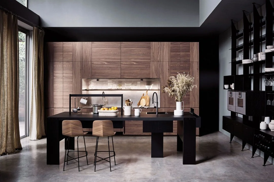 Modern kitchen with wooden cabinetry, black kitchen island, and black shelving unit, illuminated by natural light from large windows with gold curtains, decorated with vases and kitchenware.