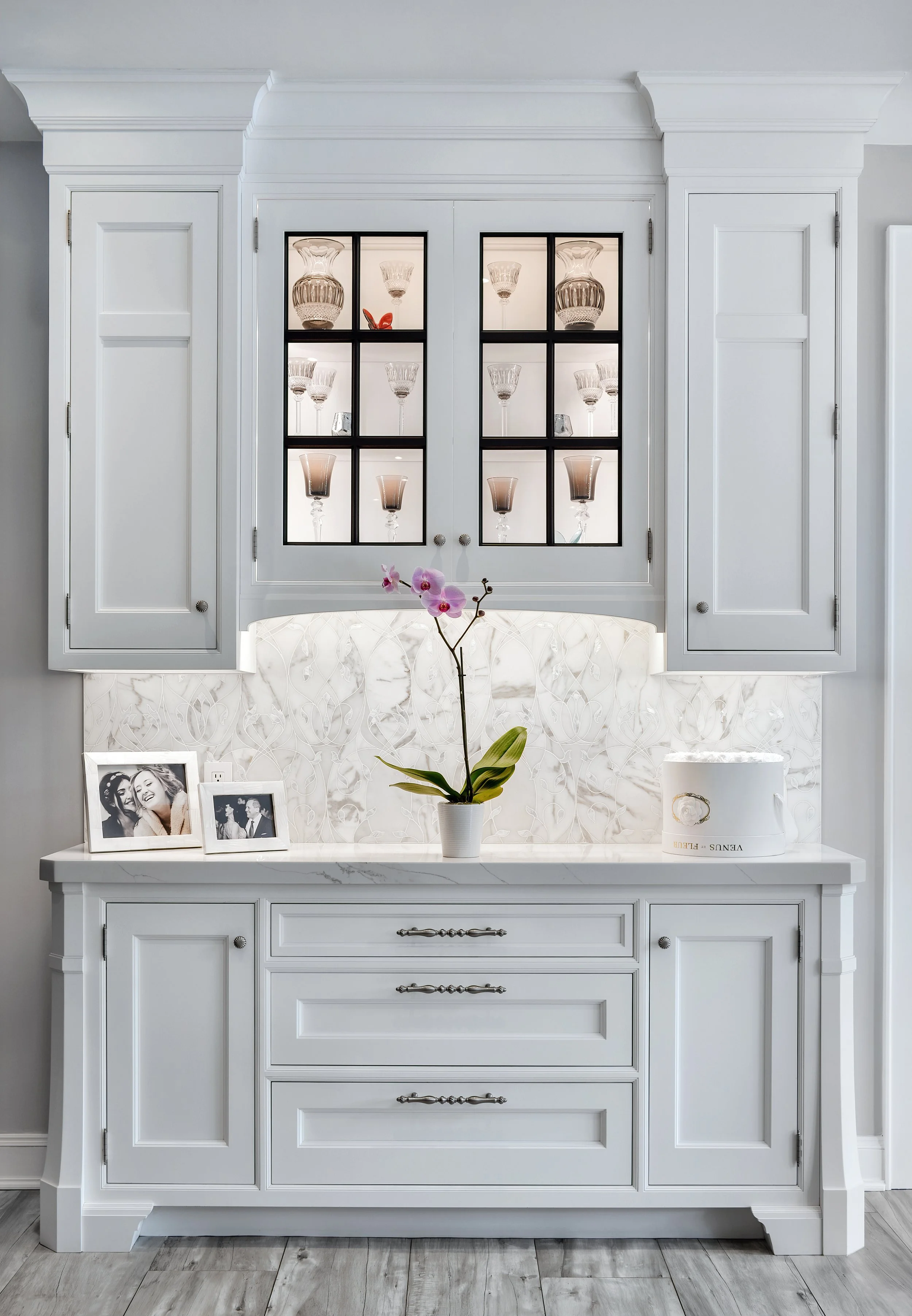 White cabinet with glassware displayed behind glass doors, decorative orchid in a white pot, framed photographs, and a cake box on a white countertop against a textured white backsplash.