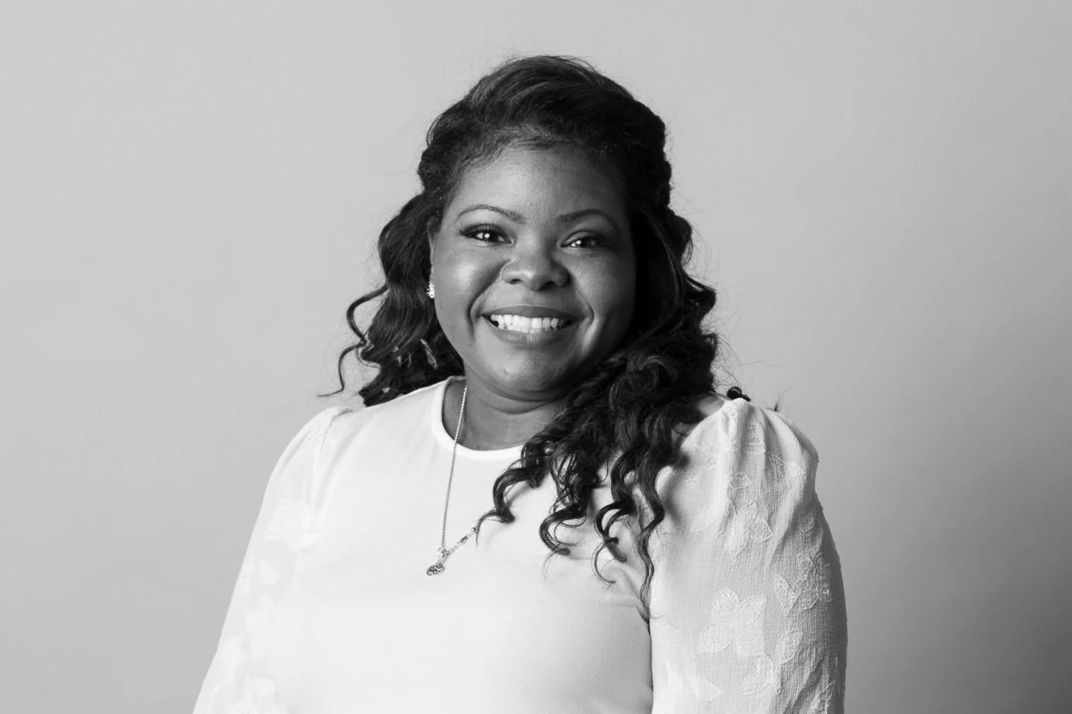 A smiling African American woman with long curly hair, wearing a light-colored top with lace details on the sleeves, a necklace with a pendant, and earrings, against a plain background.