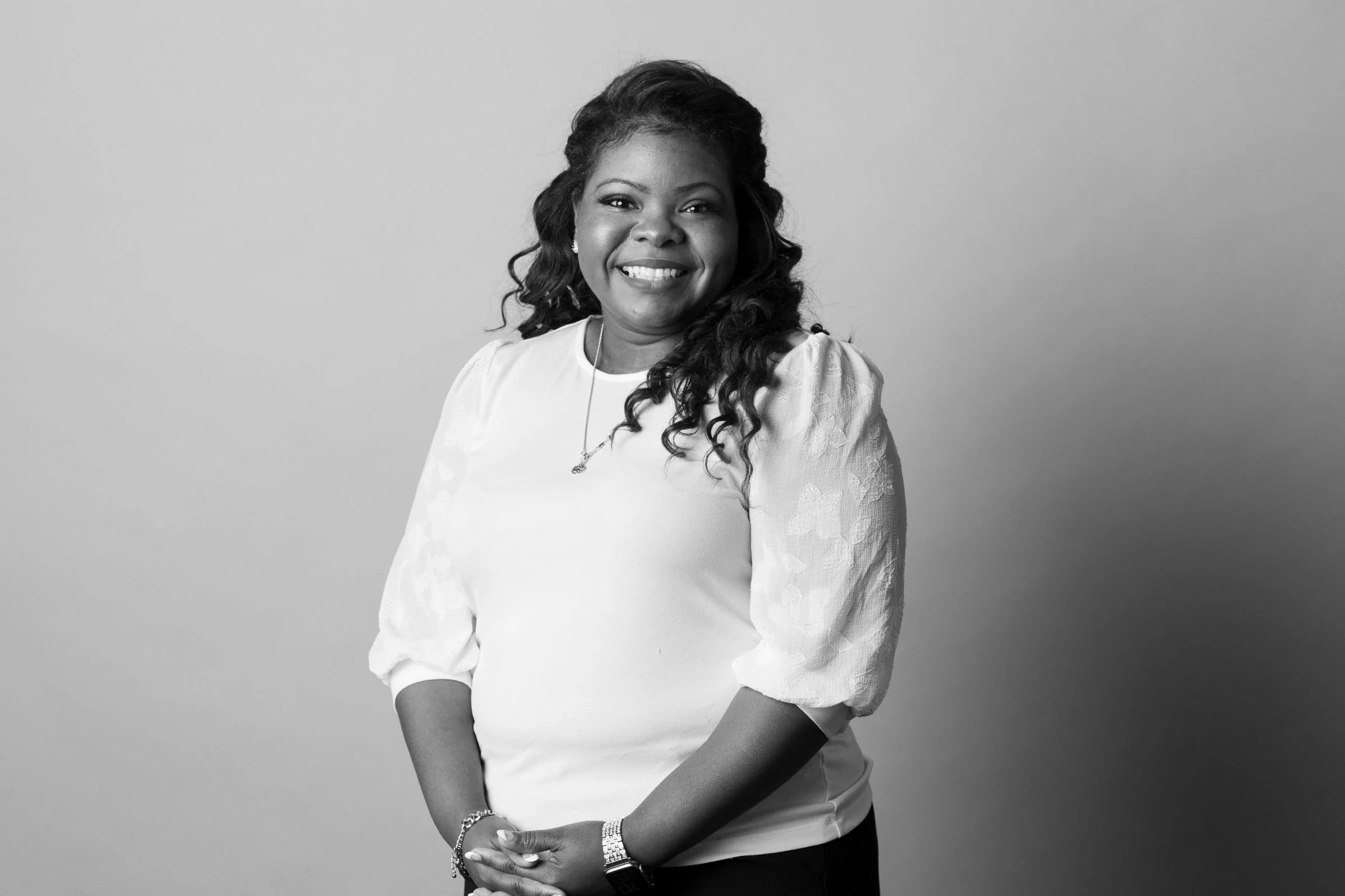 Black and white portrait photograph of a smiling woman with long wavy hair, wearing a white blouse with textured sleeves, earrings, and a watch, standing against a plain background.