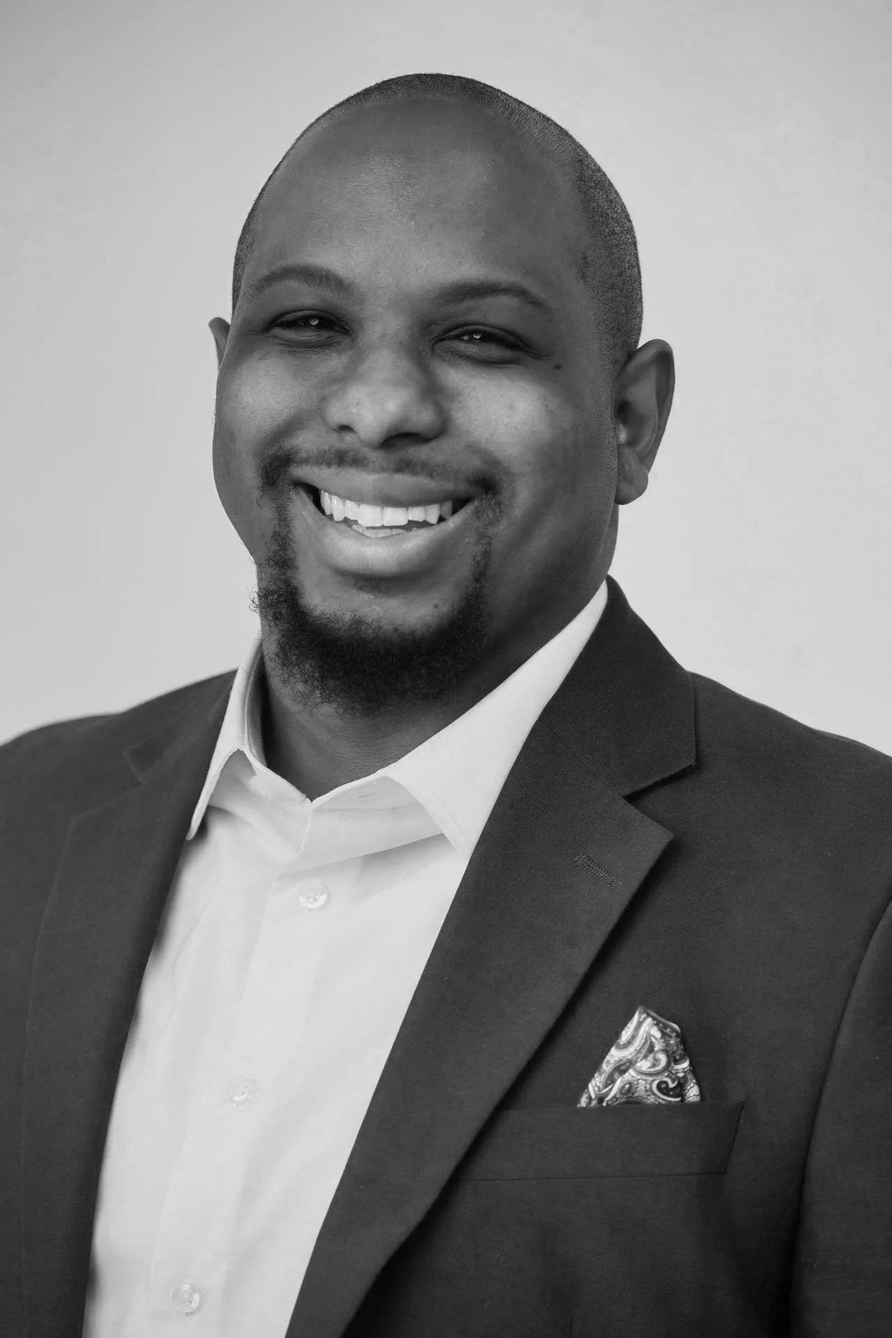 Black and white professional headshot of a smiling man in a suit and white shirt with a patterned pocket square, against a plain background.
