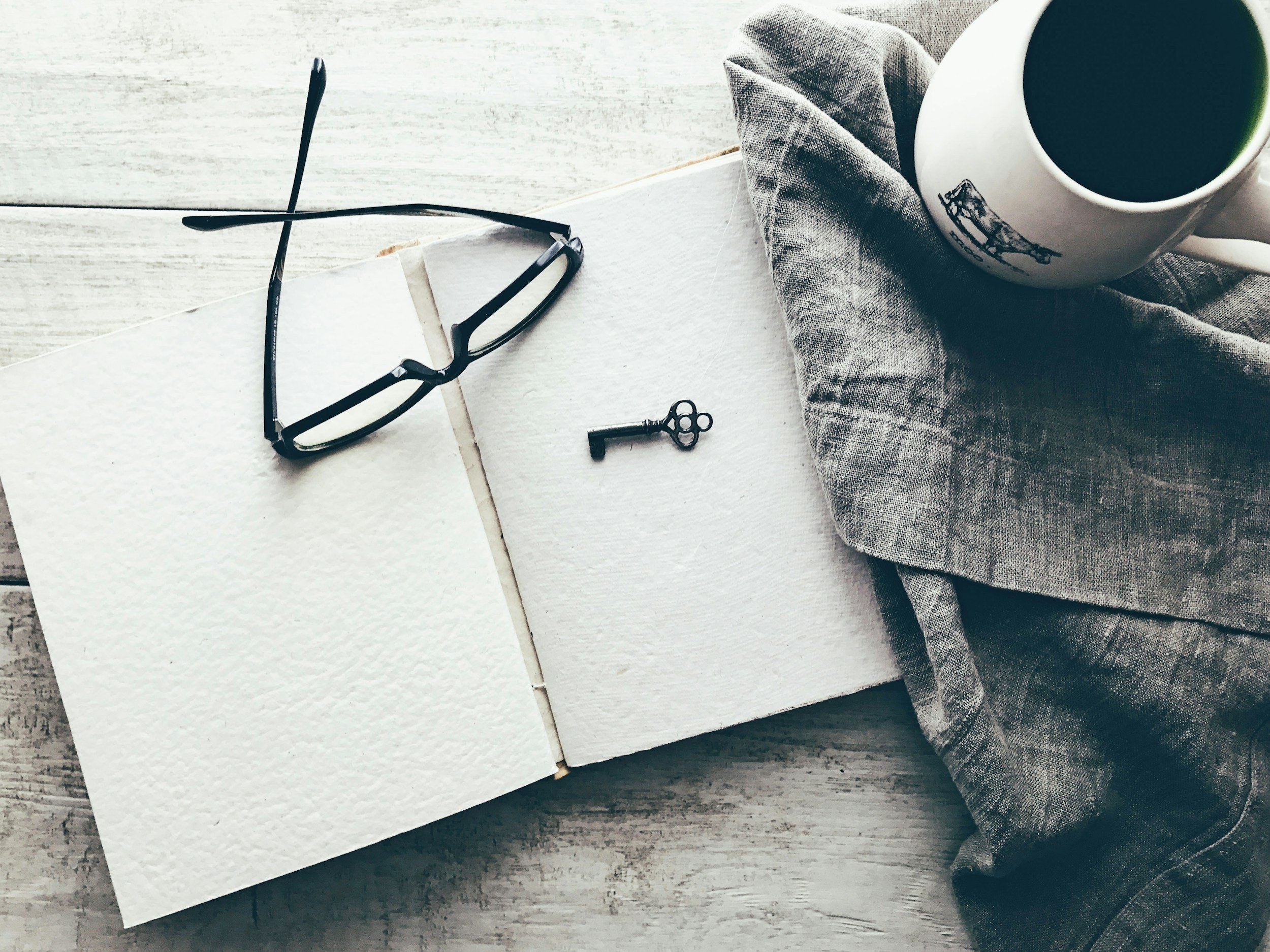 Open blank notebook with black-framed glasses, a small key, a coffee mug, and a gray cloth on a wooden surface.