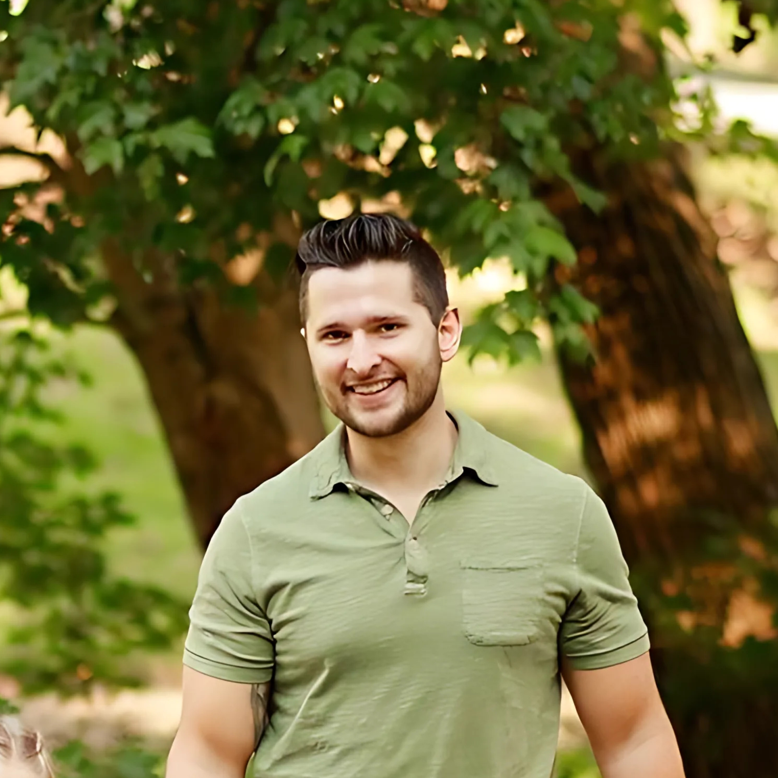A young man with dark hair styled back, smiling and wearing a light green polo shirt, outdoors in front of a large tree with green leaves.