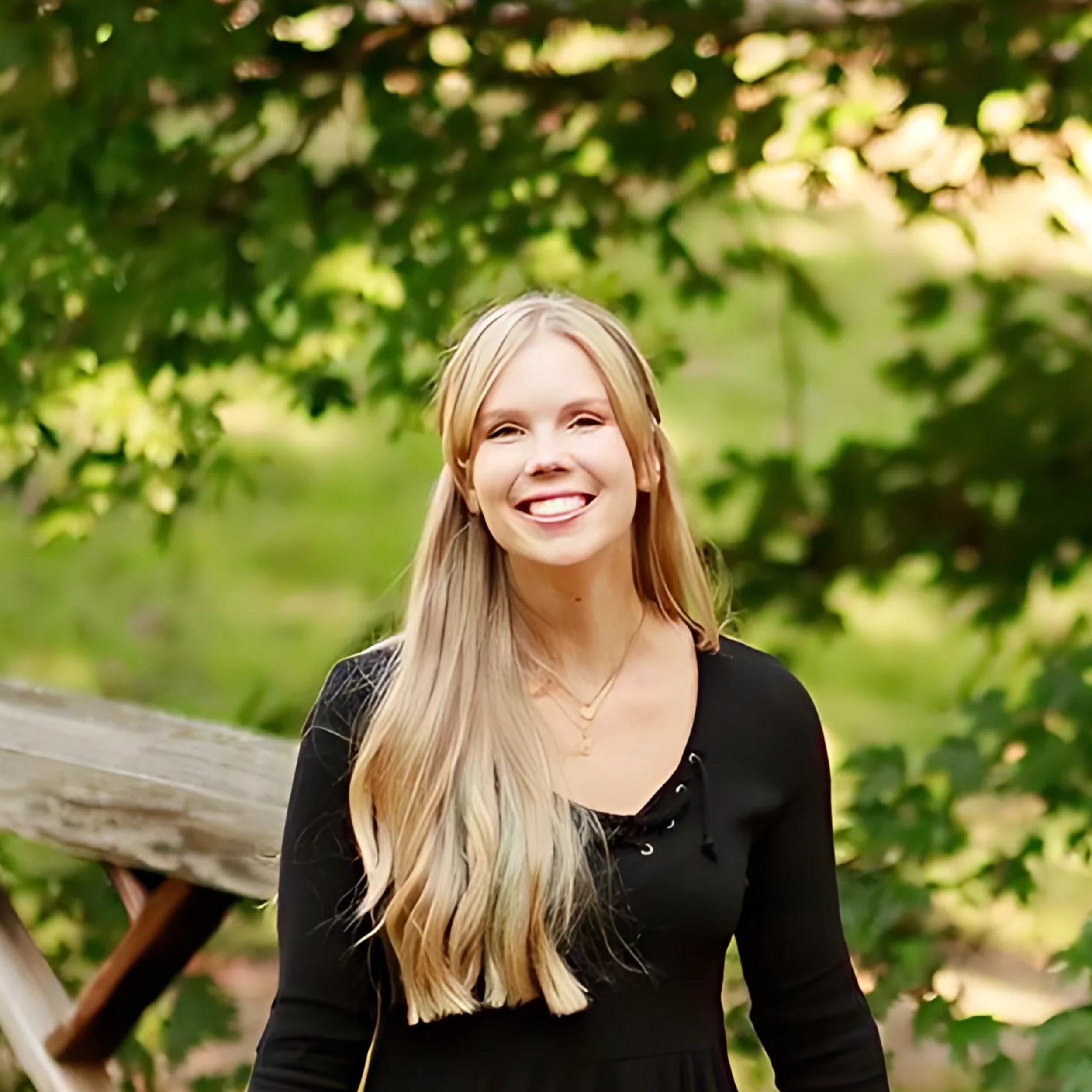 A young woman with long blonde hair wearing a black top, smiling outdoors in front of green trees and foliage.
