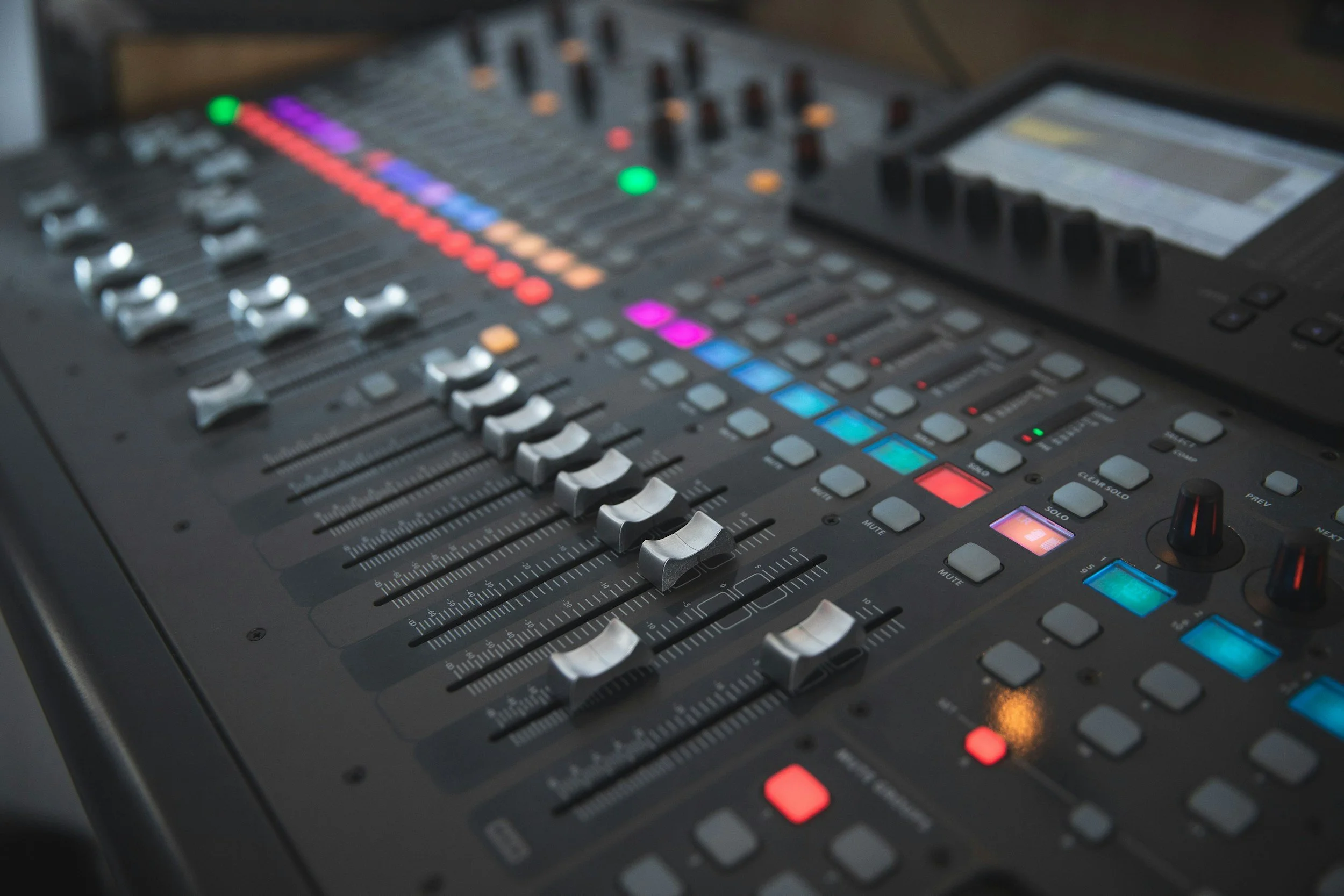 A professional audio mixing console with various controls, sliders, and colorful LED indicators in a recording studio.
