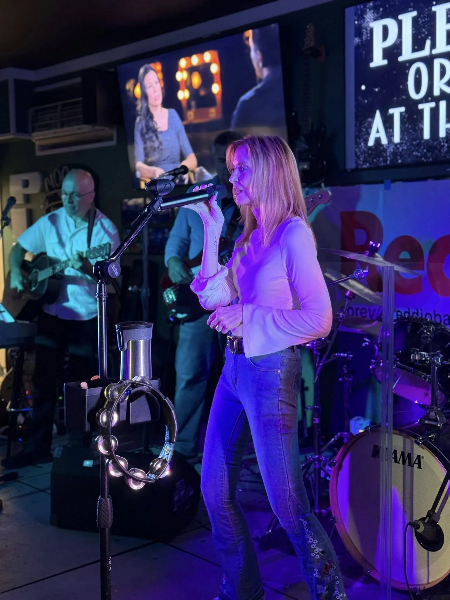 A woman singing into a microphone on stage with a band playing behind her, illuminated by colorful stage lights.