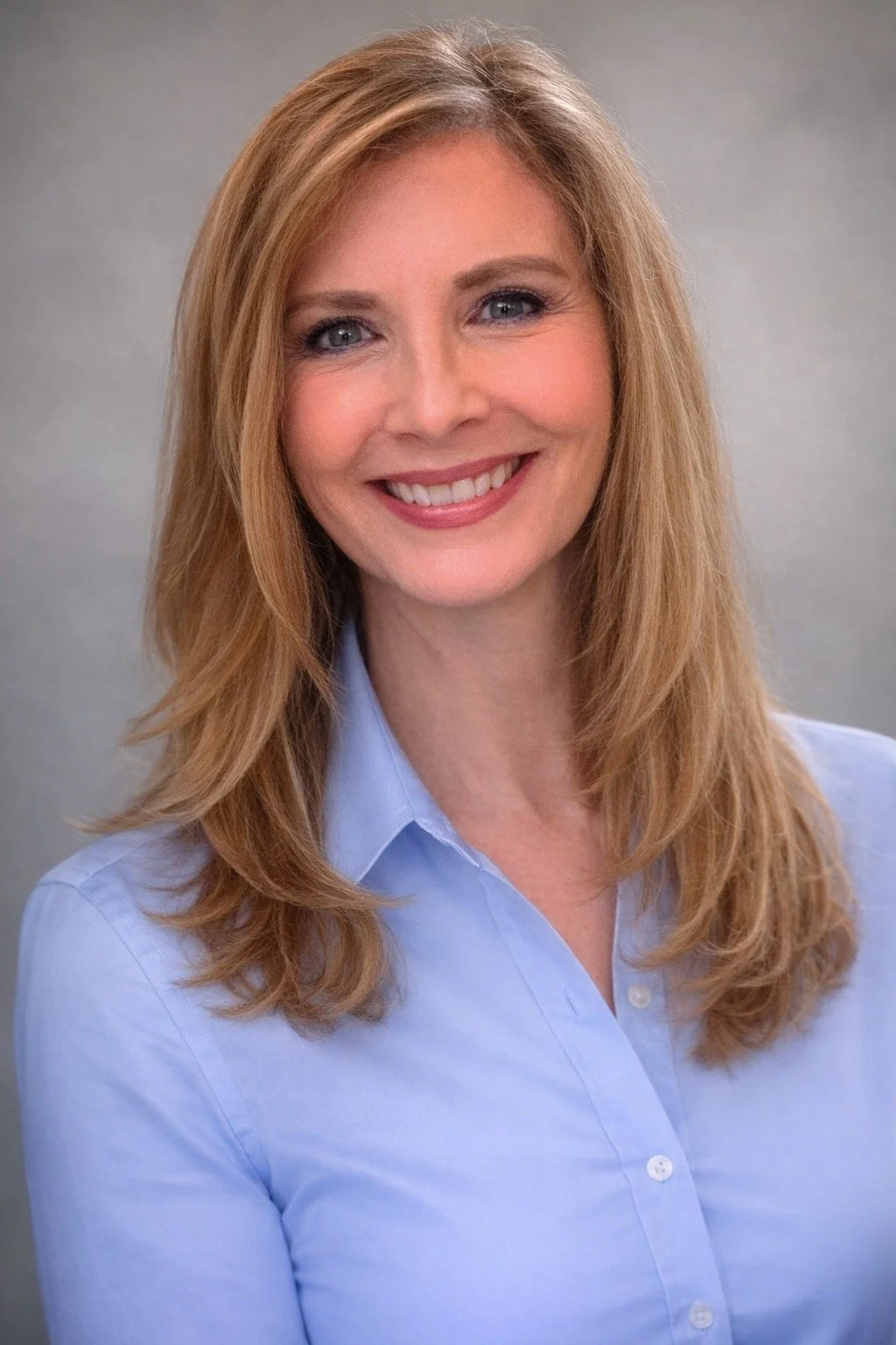 A woman with long blonde hair, blue eyes, and a bright smile, wearing a light blue button-up shirt against a soft gray background.