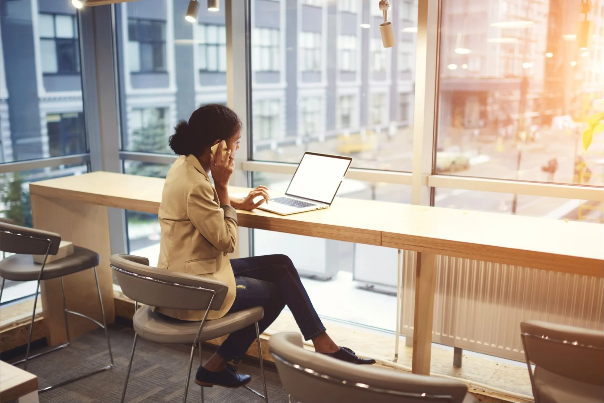 A concierge sitting at a high wooden counter by a large window, talking on a cellphone, with a laptop open in front of her, in a modern office or cafe with city buildings outside.