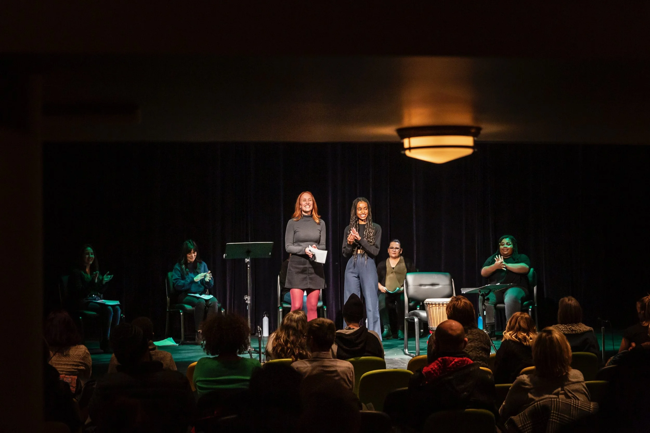 Two women stand on stage in front of an audience, with four women seated behind them. The stage is dark with a black curtain background, and the audience is sitting in chairs watching the presentation.