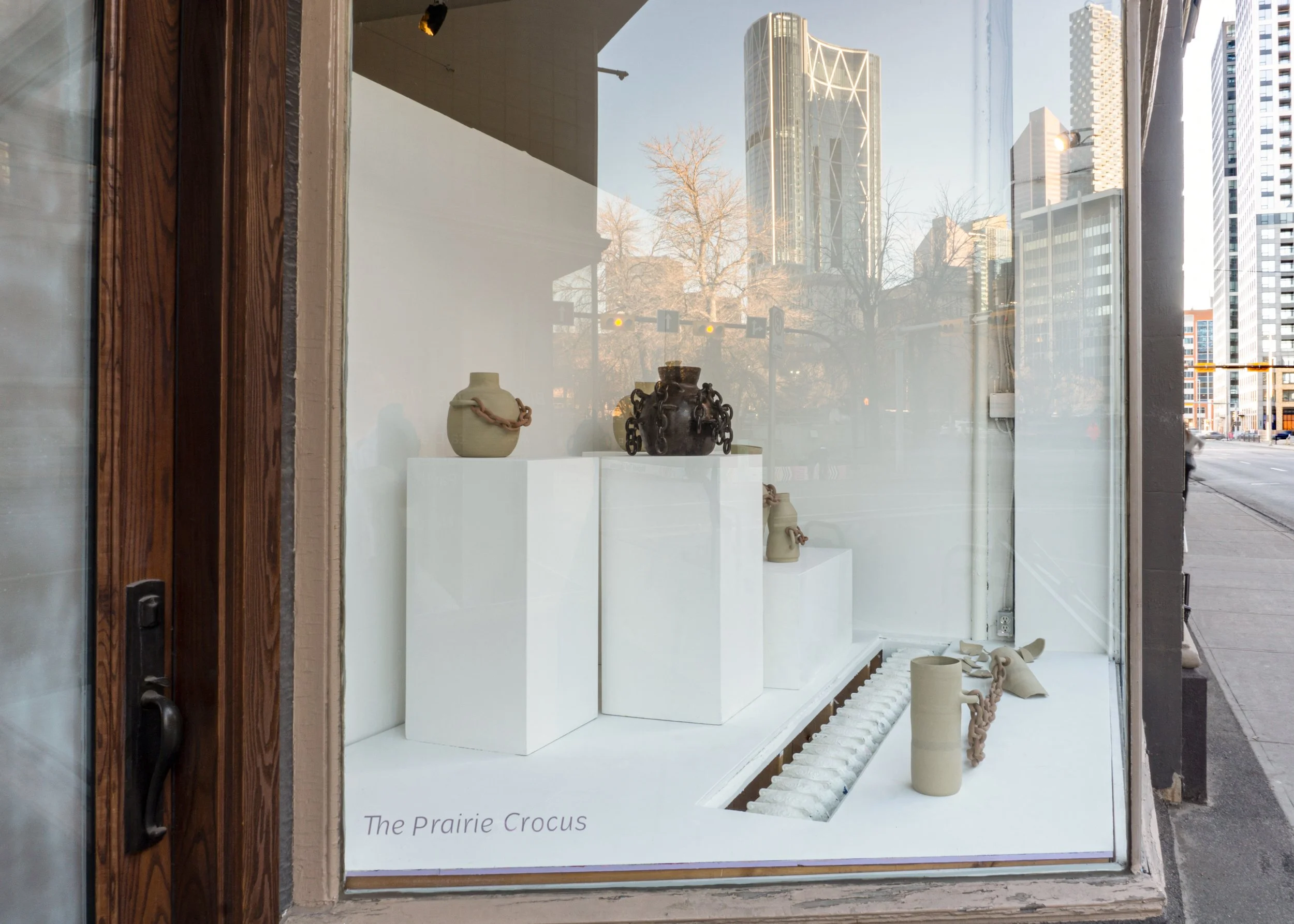 Art display of ceramic vases and sculptures in a gallery window with cityscape reflection.