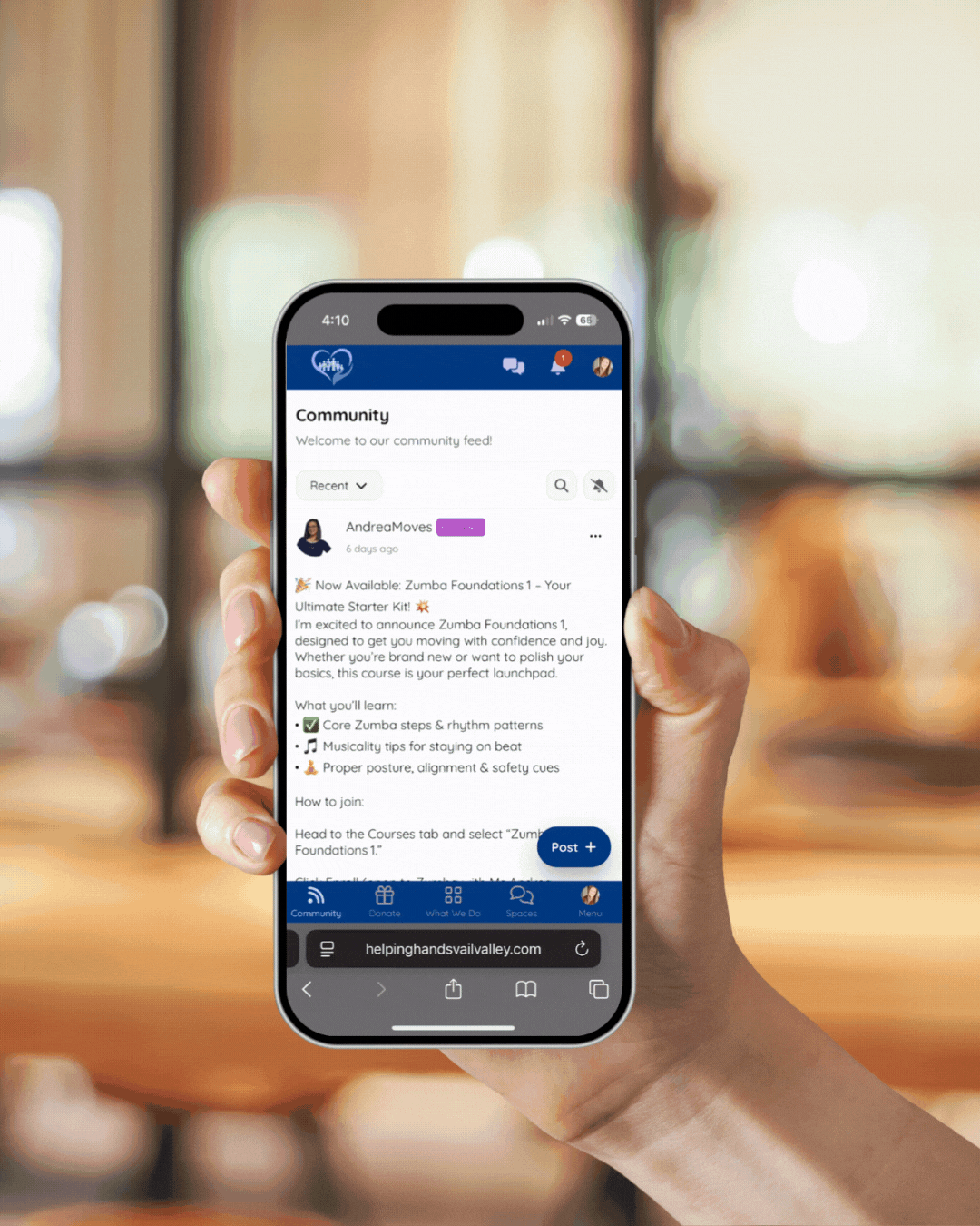 A person holding a smartphone displaying a community social media feed with a post about Zumba Foundations 1 class.