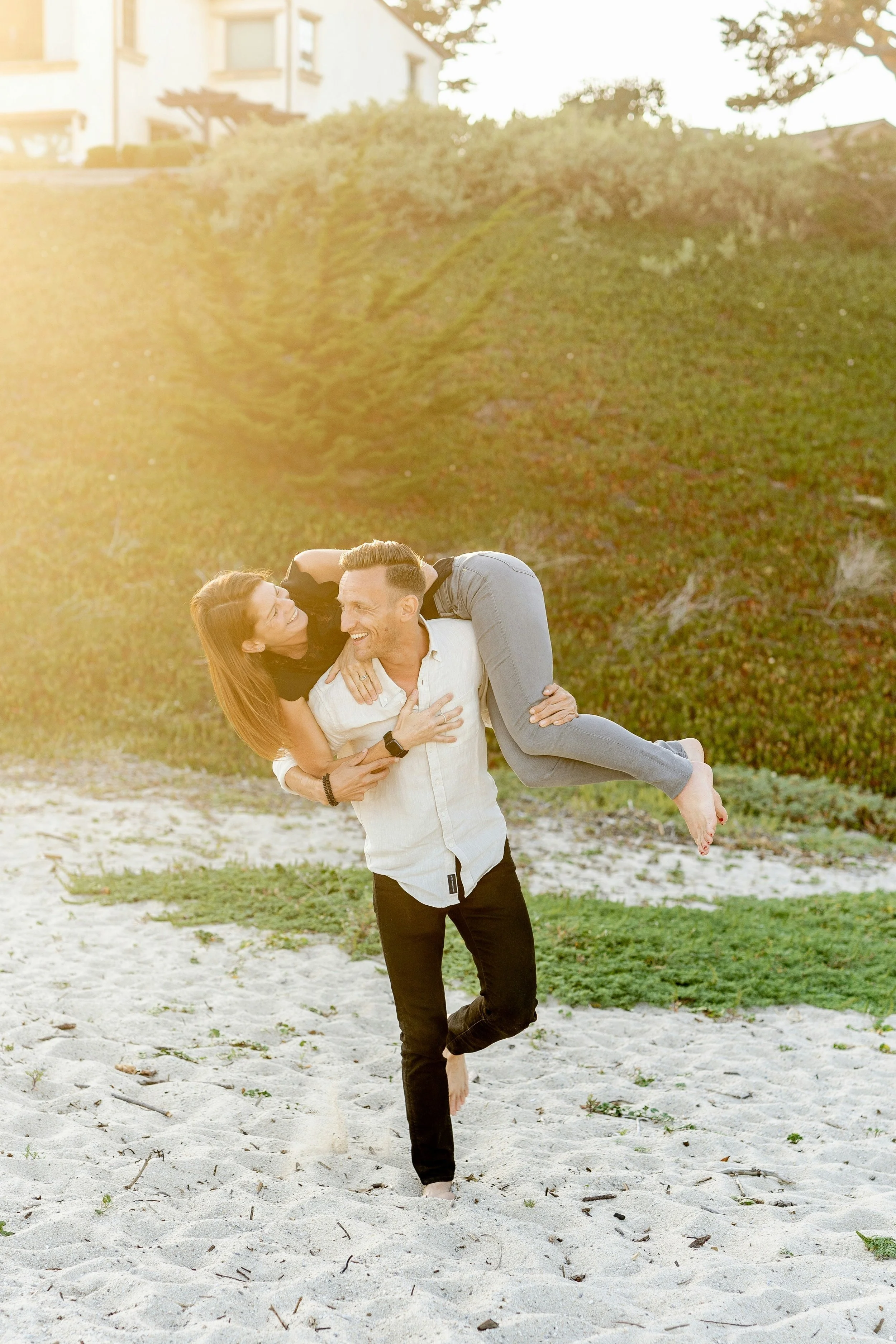 A man is holding a woman on his shoulders while walking barefoot on a sandy beach in the late afternoon or early evening. They are smiling and enjoying each other's company, with green bushes and a house visible in the background.