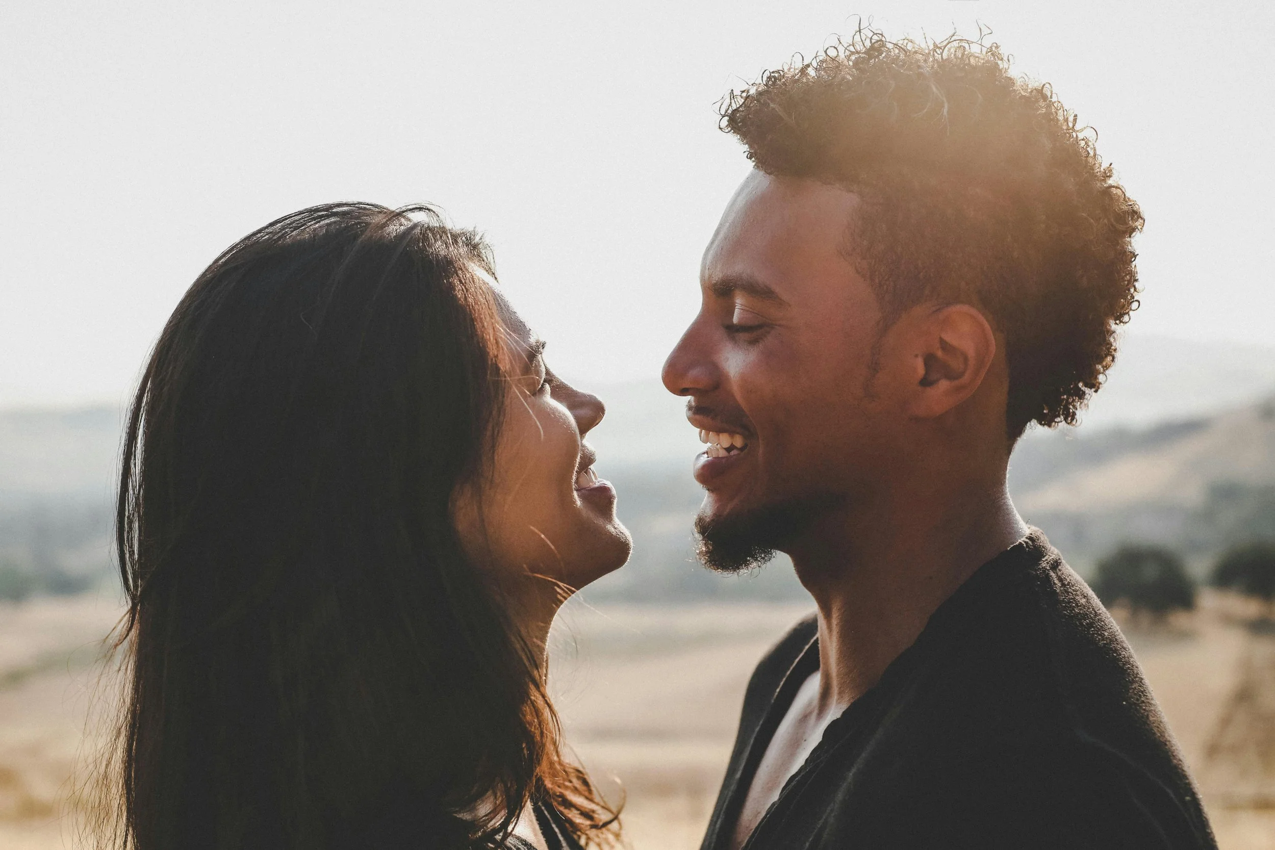 A man and woman smiling and close to each other outdoors during daytime with natural background.
