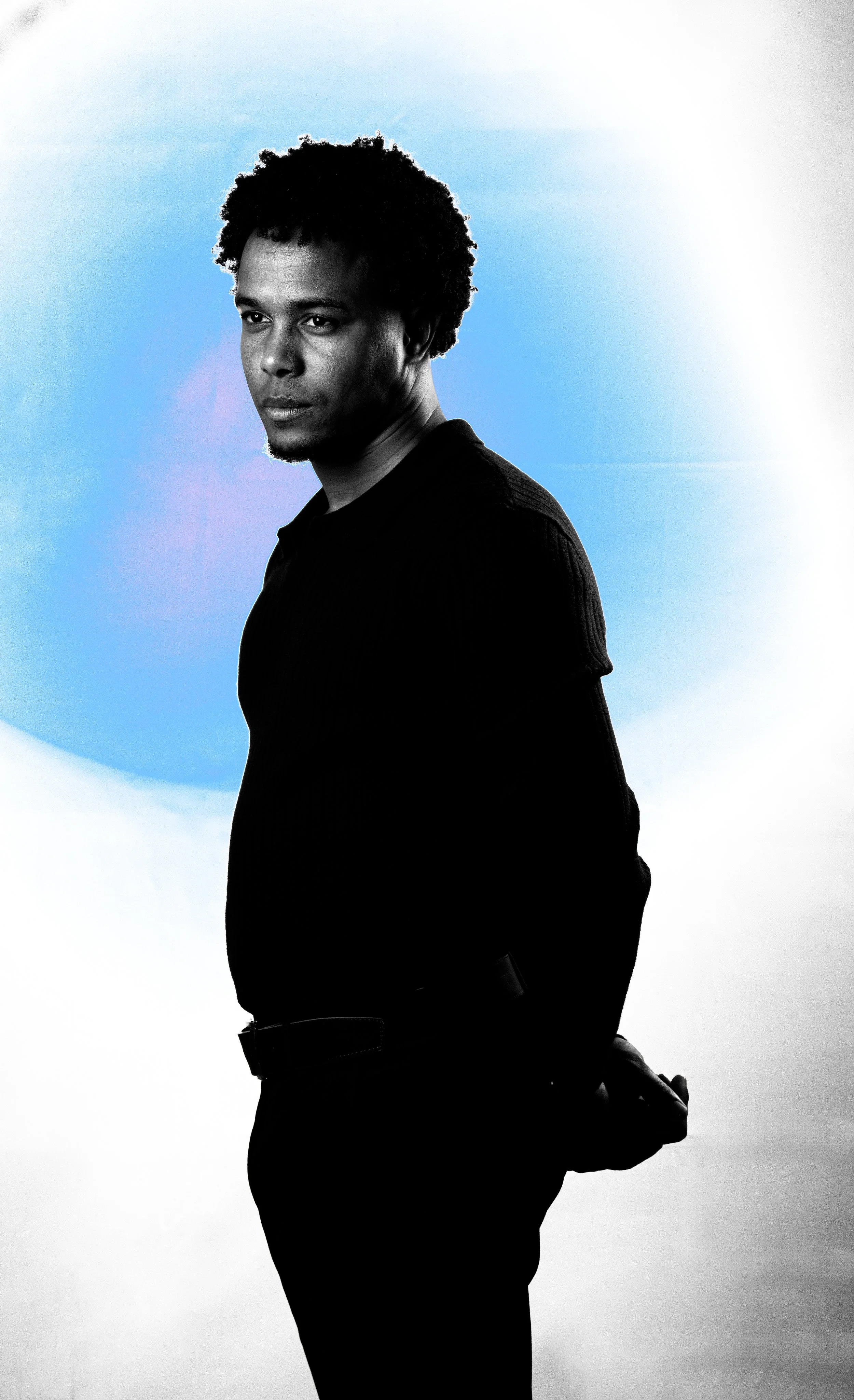 Ritratto di un uomo con capelli ricci neri, abbigliamento nero, sfondo sfumato con sfumature blu e bianco