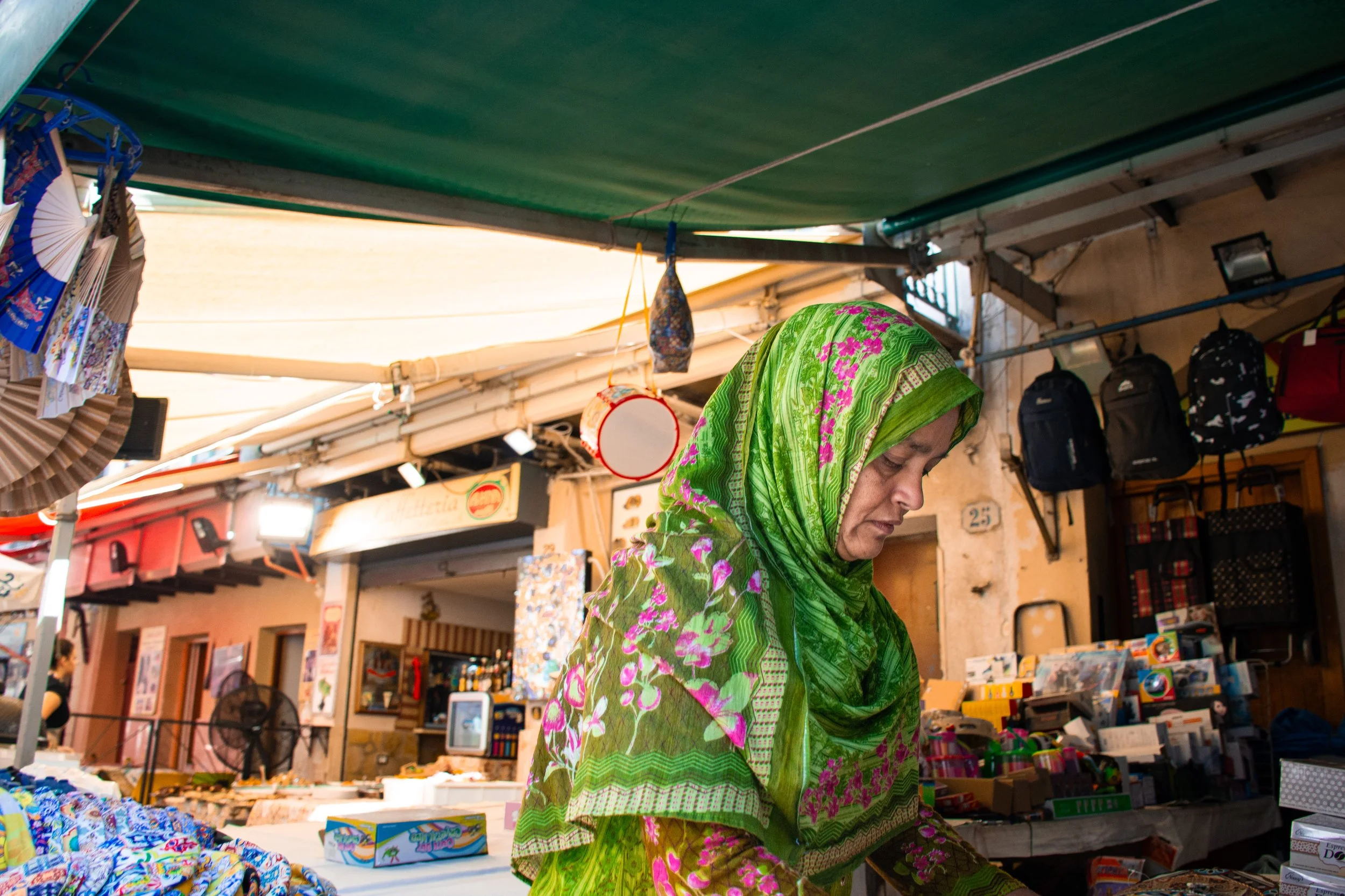 Una donna indossa un hijab verde con motivi floreali mentre fa acquisti in un mercato all'aperto, con bancarelle piene di vari prodotti in vendita.