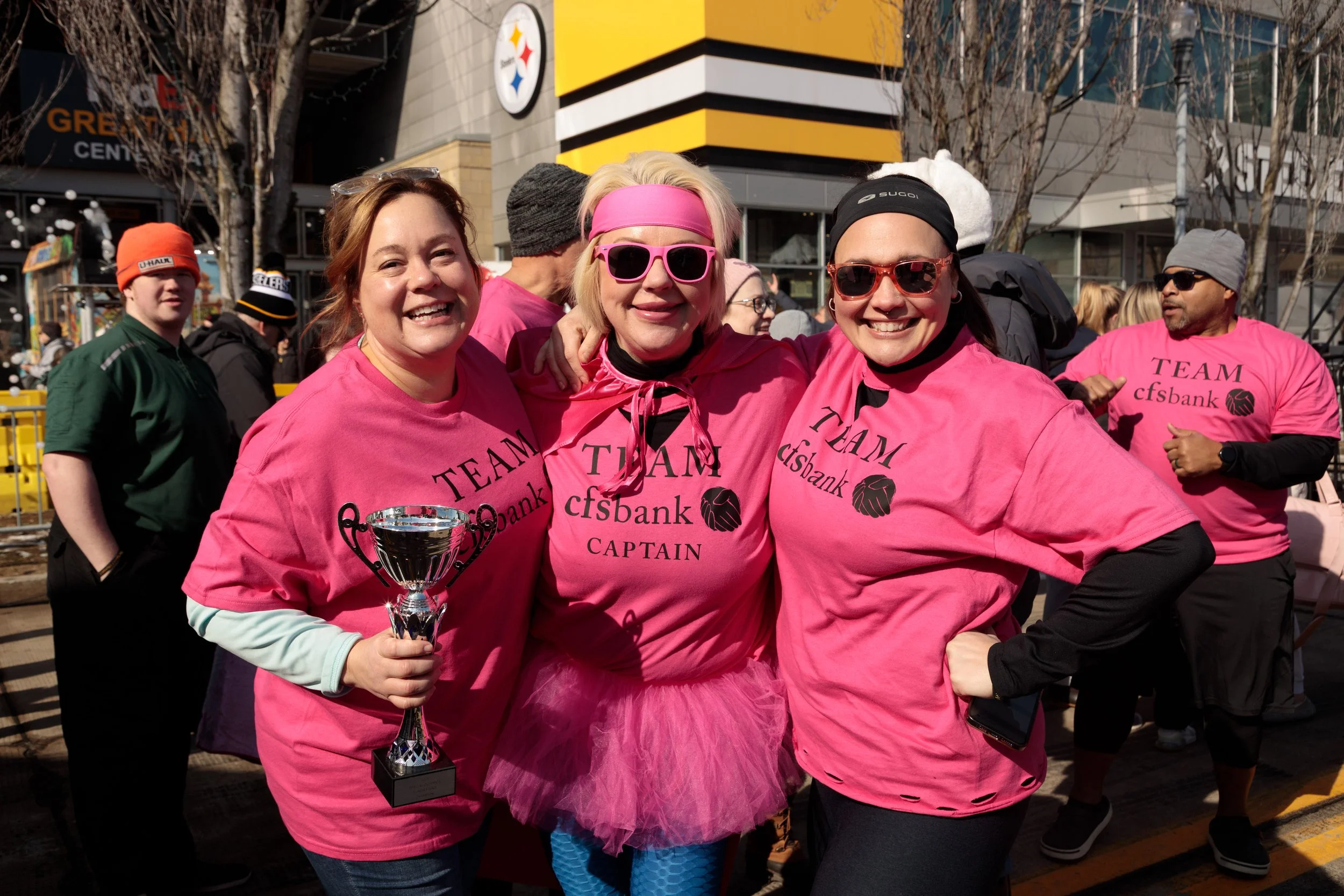 Group of women wearing pink t-shirts, some with "TEAM cfsbank" and "CAPTAIN" written on them, celebrating outdoors on a sunny day at a charity event or race, with some smiling and holding a trophy.