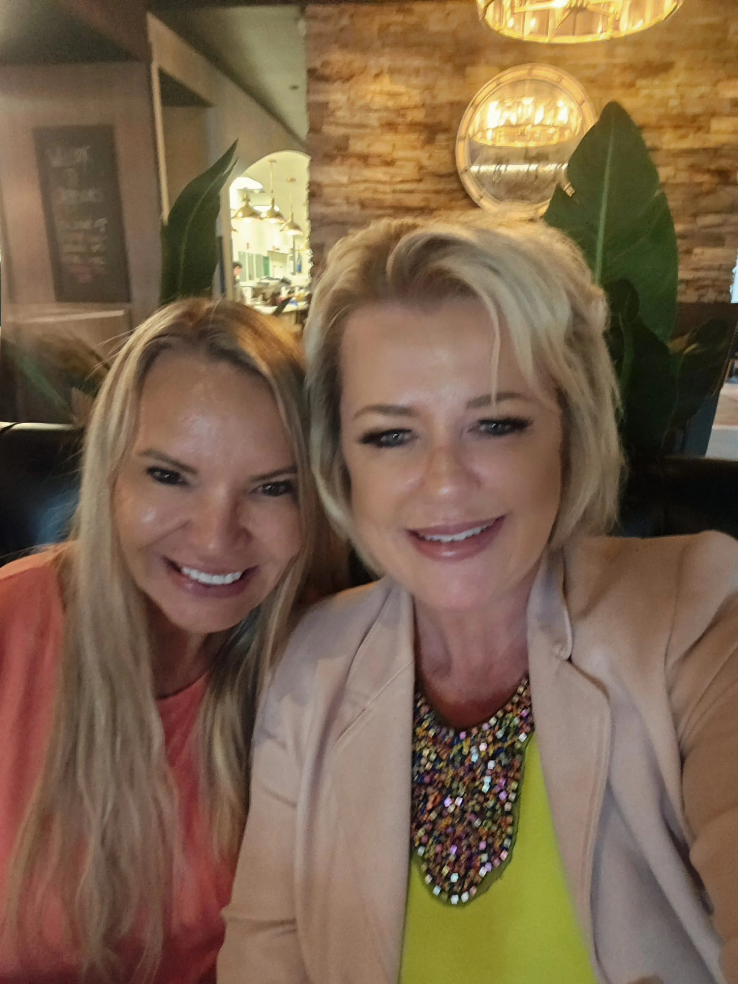 Two women taking a selfie at a restaurant, smiling, with a warm, cozy interior featuring a brick wall, plants, and warm lighting.