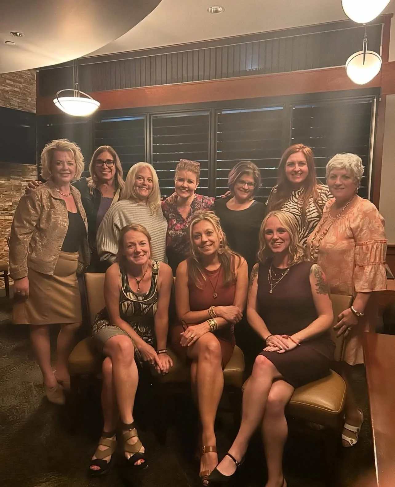 A group of ten women gathered in a restaurant or lounge, some sitting on a cushioned bench and others standing behind. They are smiling and dressed in semi-formal attire, with a wooden and brick interior and modern lighting fixtures overhead.