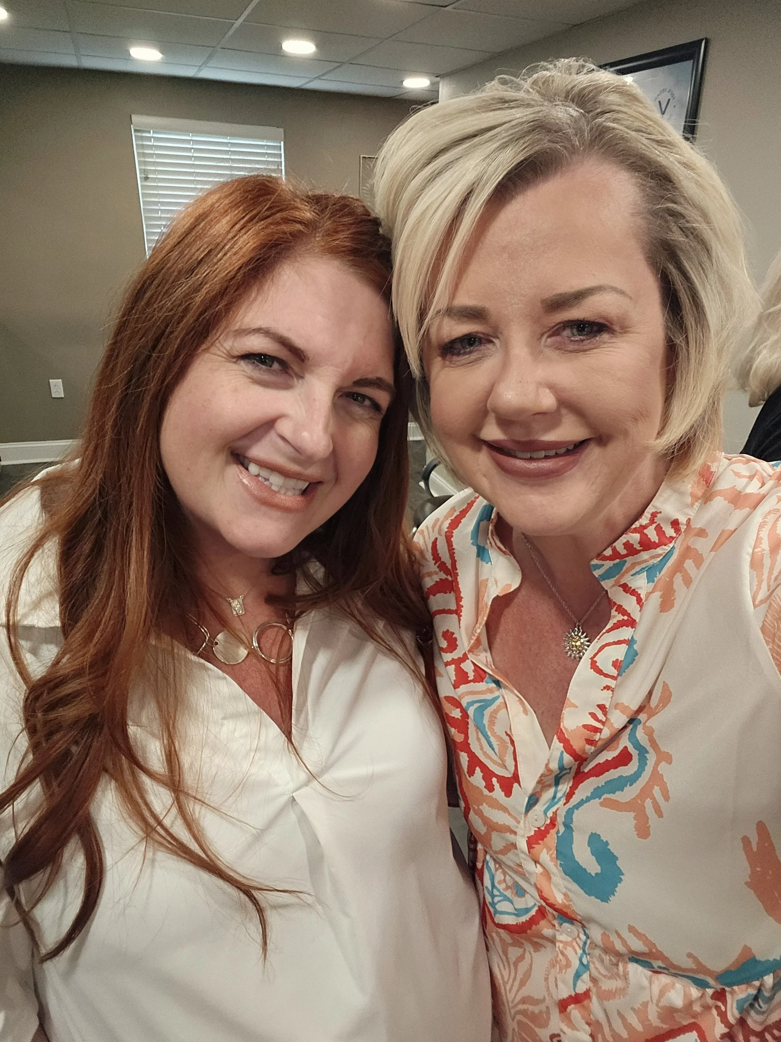 Two women smiling and taking a selfie inside a room with gray walls, a window with blinds, and a ceiling with lights.