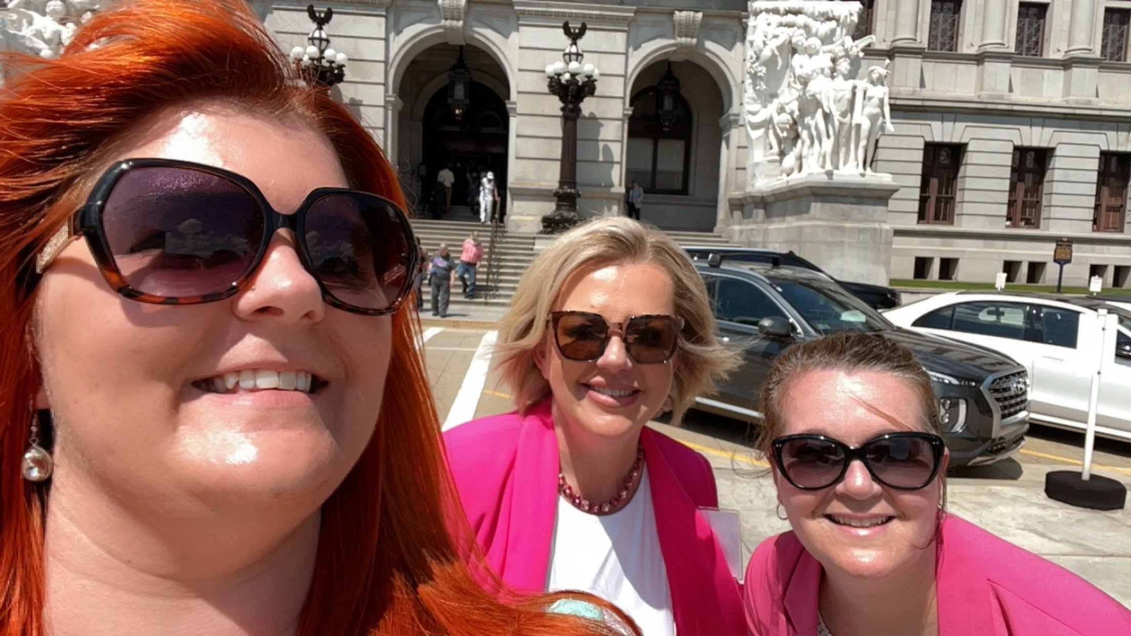 Three women wearing sunglasses and pink clothing taking a selfie outside a large, ornate building with stone sculptures and decorative lights.