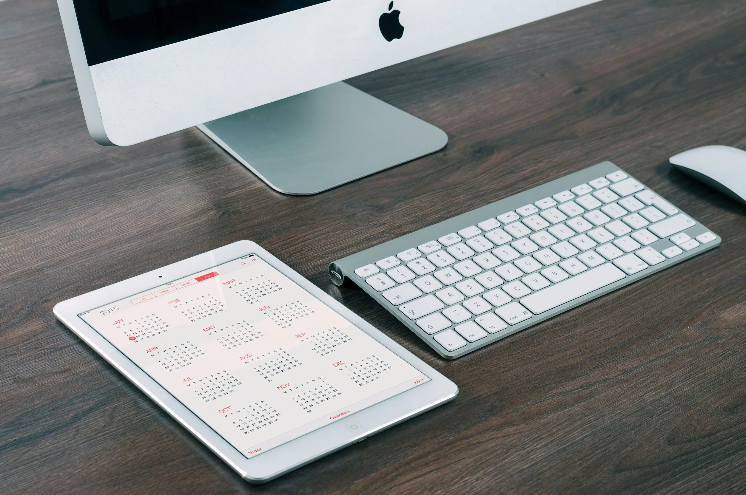 Apple iMac computer, wireless keyboard, mouse, and tablet displaying a yearly calendar on a wooden desk.