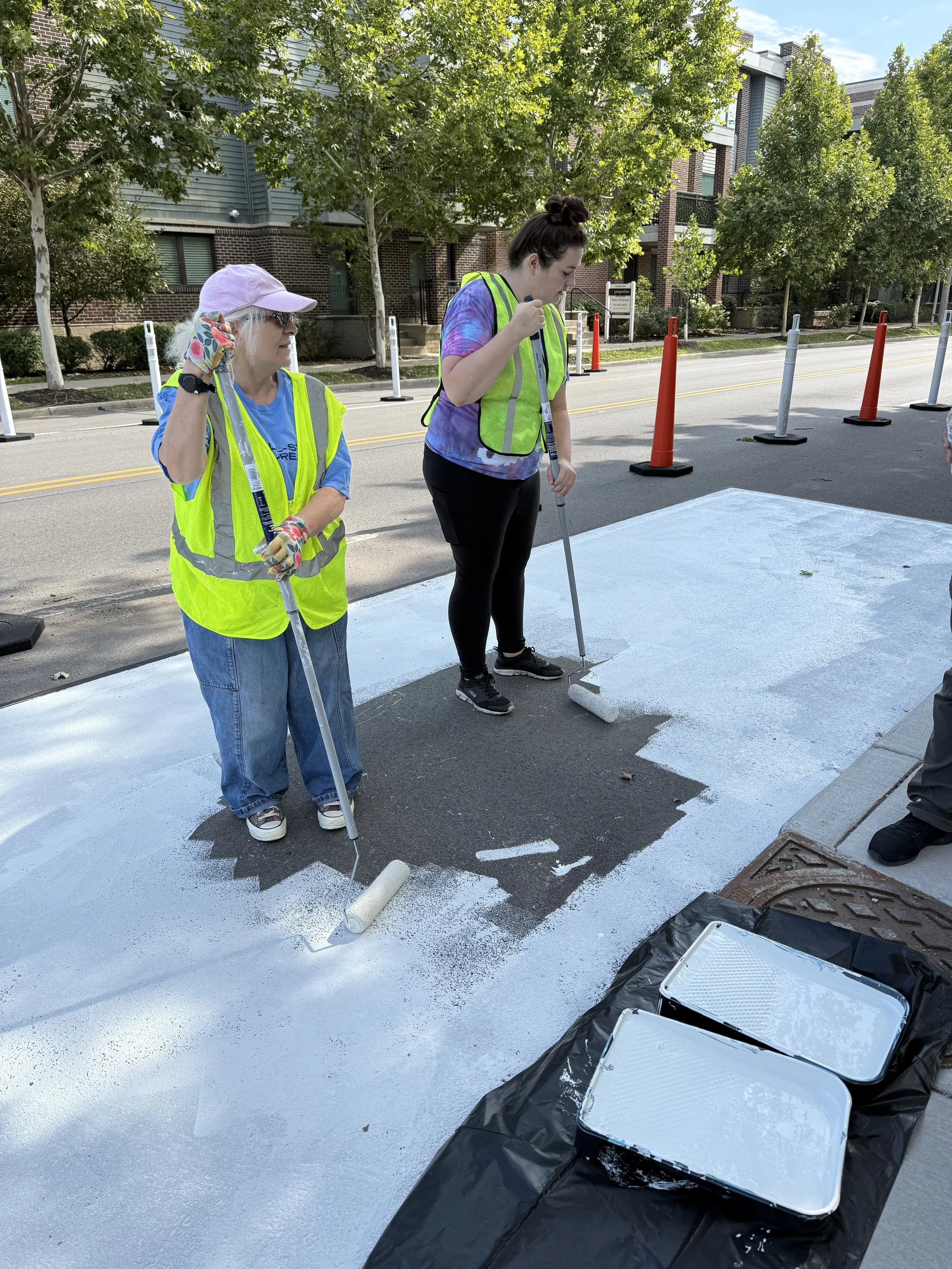 Two women in safety vests painting a crosswalk on the street with white paint, while a person off-camera paints next to them. There are orange and white traffic cones and barriers in the background.