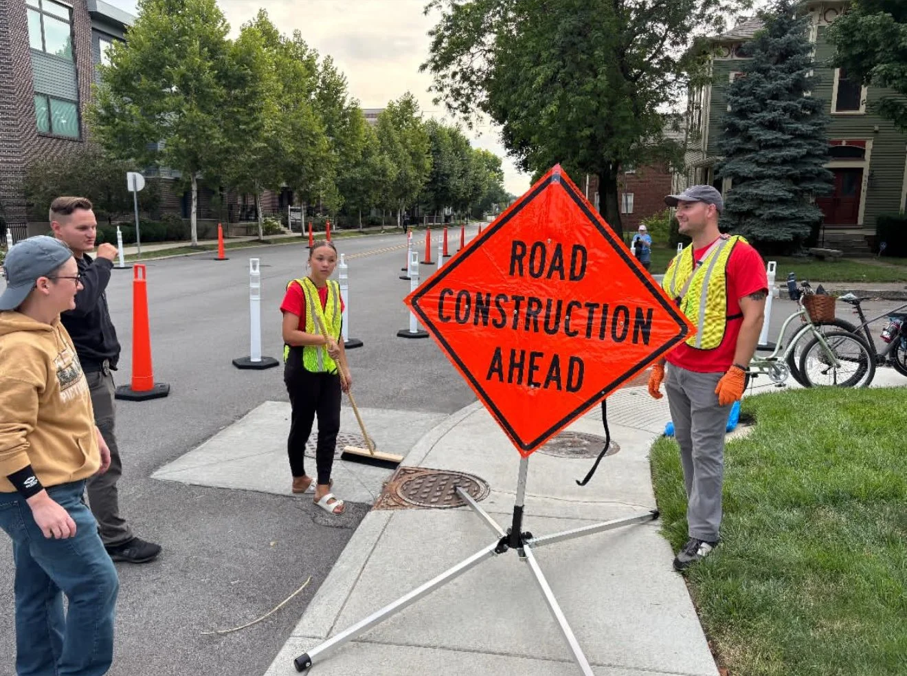 Four people standing near a bright orange 'Road Construction Ahead' sign on a city street. Two of them are wearing high-visibility vests, one person is sweeping leaves, and a fourth is talking to the group. Cones are set up along the road, and trees and houses are in the background.